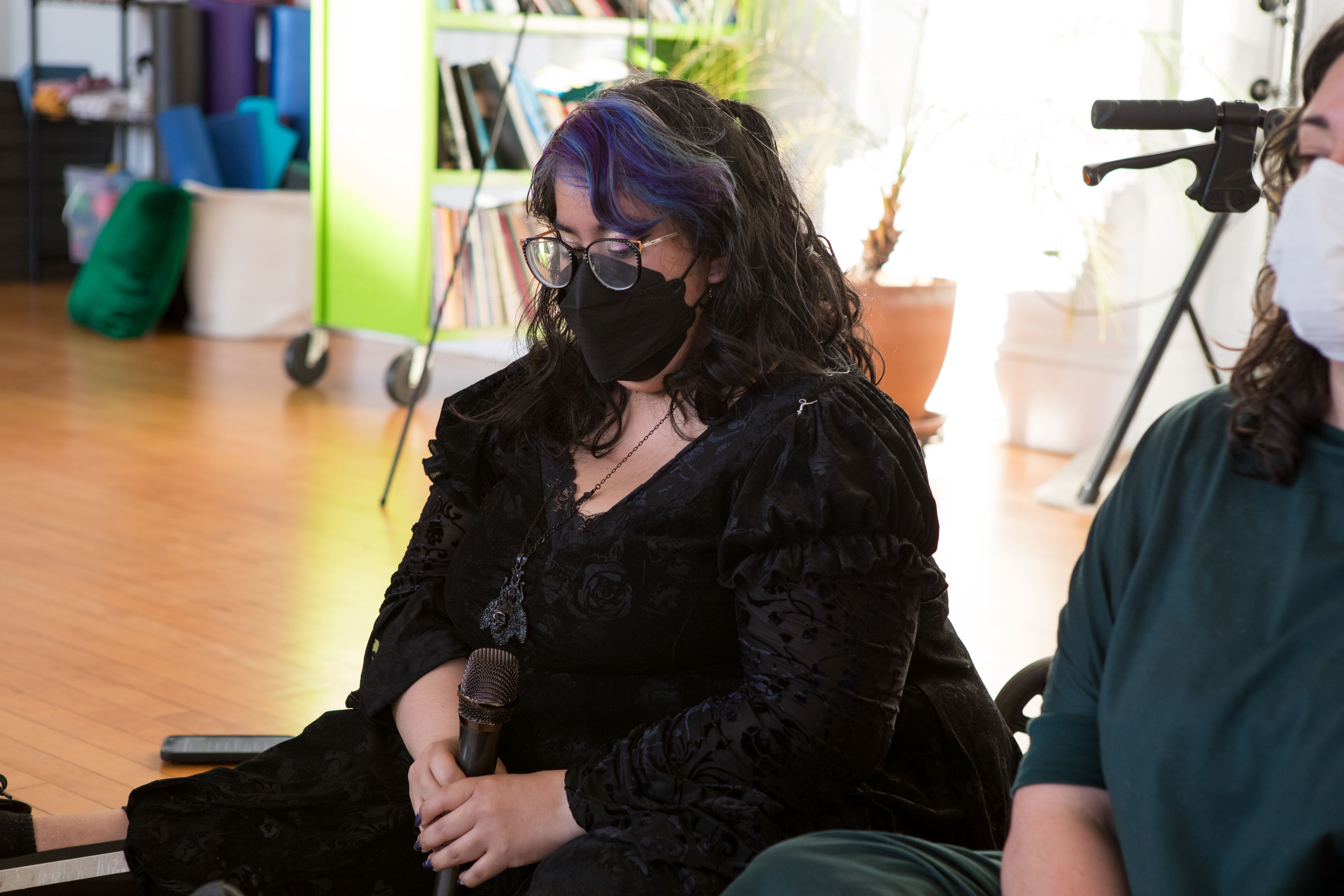 A person with dark hair with purple highlights, wearing glasses, a black mask, and a black dress, sits on a wooden floor holding a microphone. They are looking down with their eyes closed.