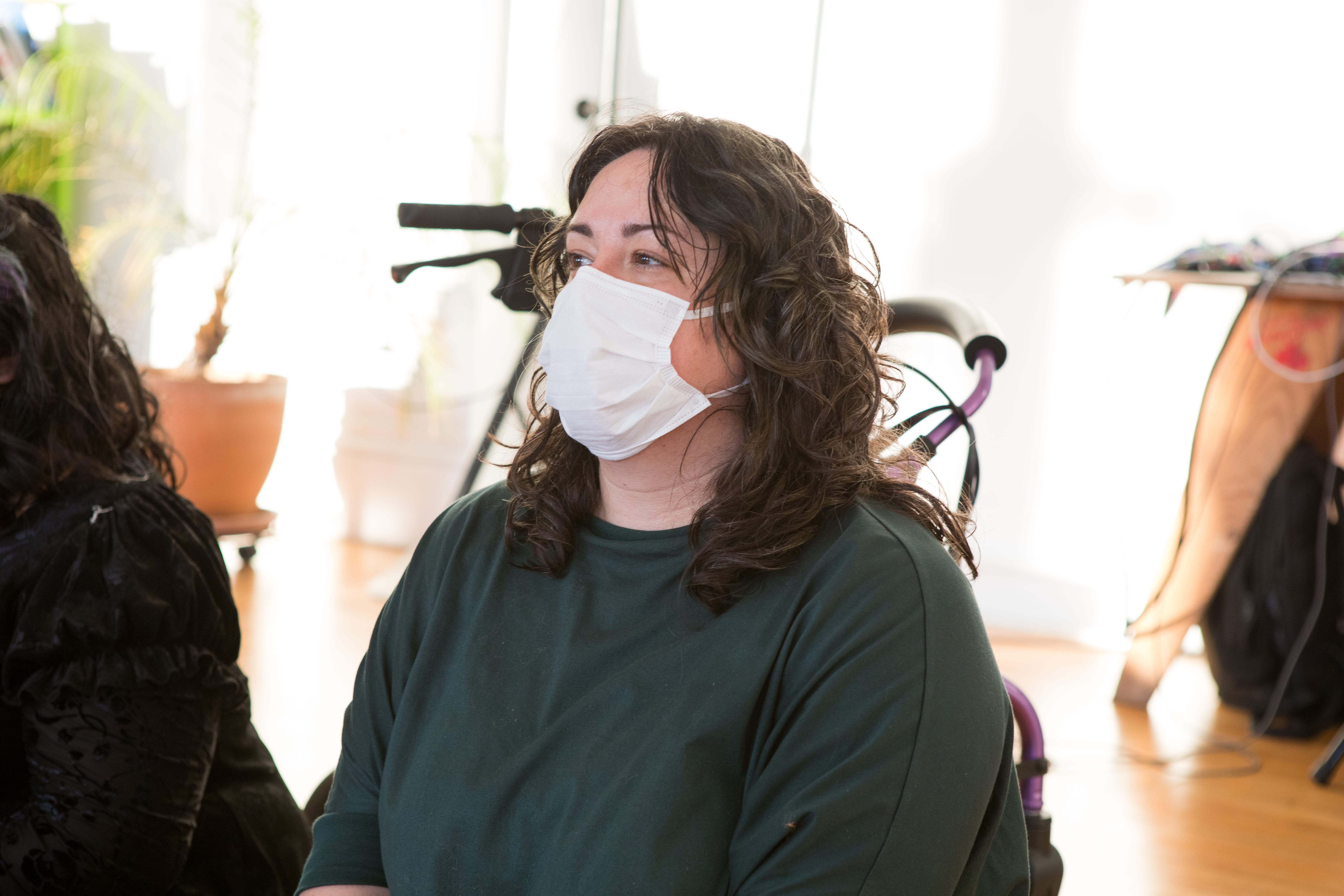 A close-up shot of a person with long, dark, curly hair, wearing a white face mask and a green shirt. They are looking off to the left of the frame, and the top of a walker is visible behind them.