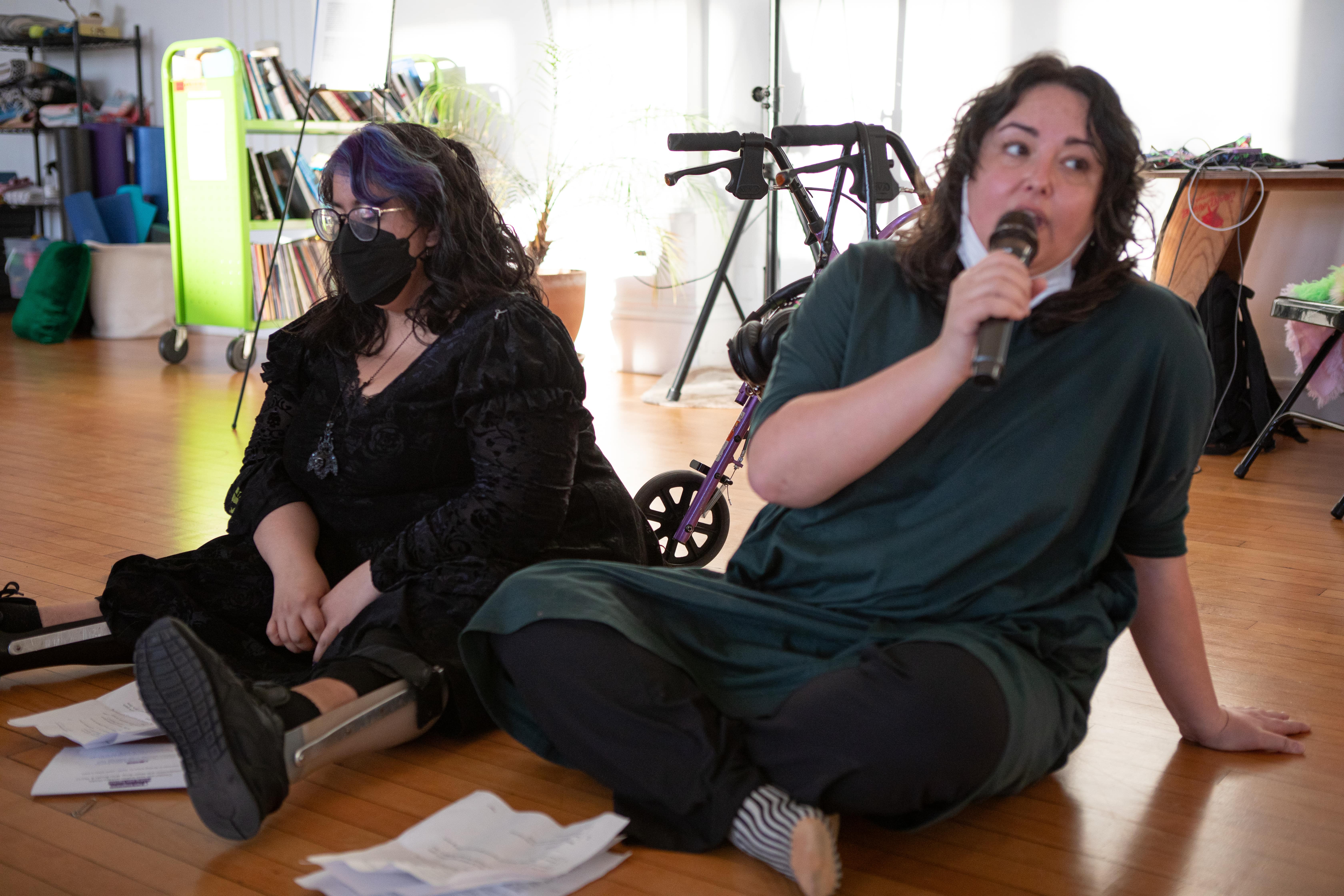 Two people are sitting on a wooden floor. The person on the right is speaking into a microphone while the person on the left, who is wearing a black dress and listens with their eyes closed. Both are wearing face masks.