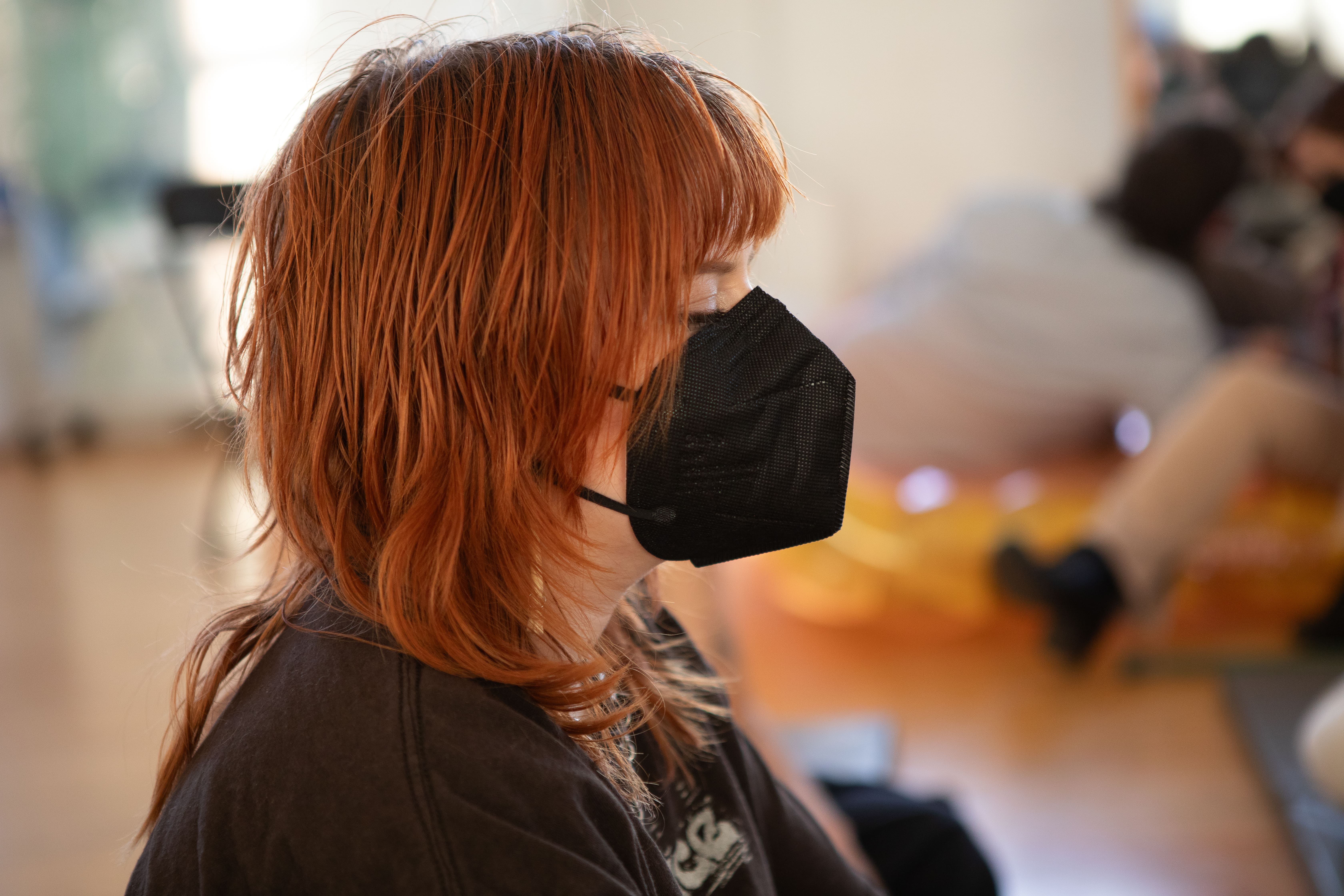 A side-profile shot of a person with short, bright red hair, wearing a black face mask and looking off to the right.