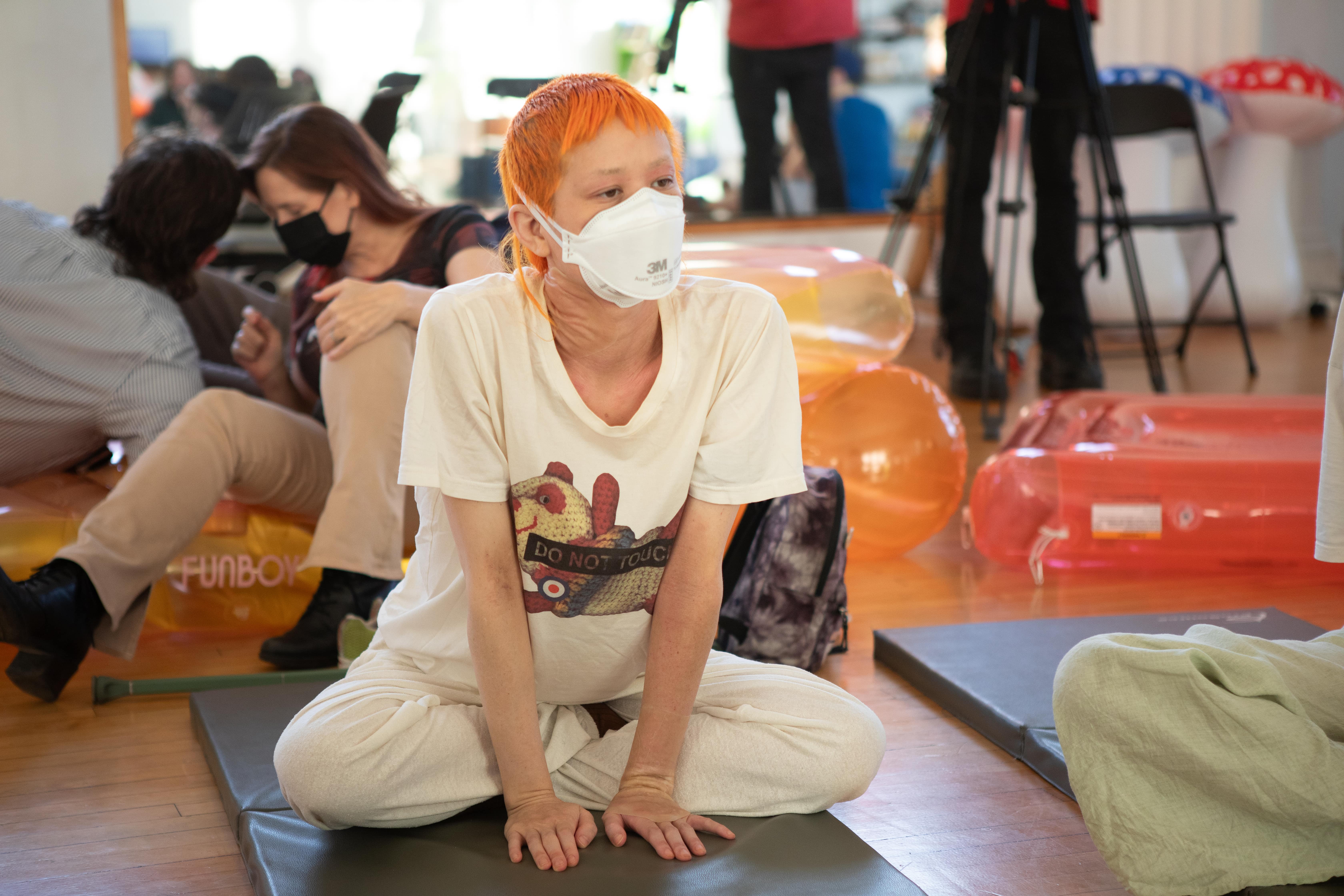 A person with short, bright orange hair and a white 3M face mask sits cross-legged on a grey mat on a wooden floor. They are wearing a light-colored t-shirt with a graphic on it. In the background, there are inflatable objects.