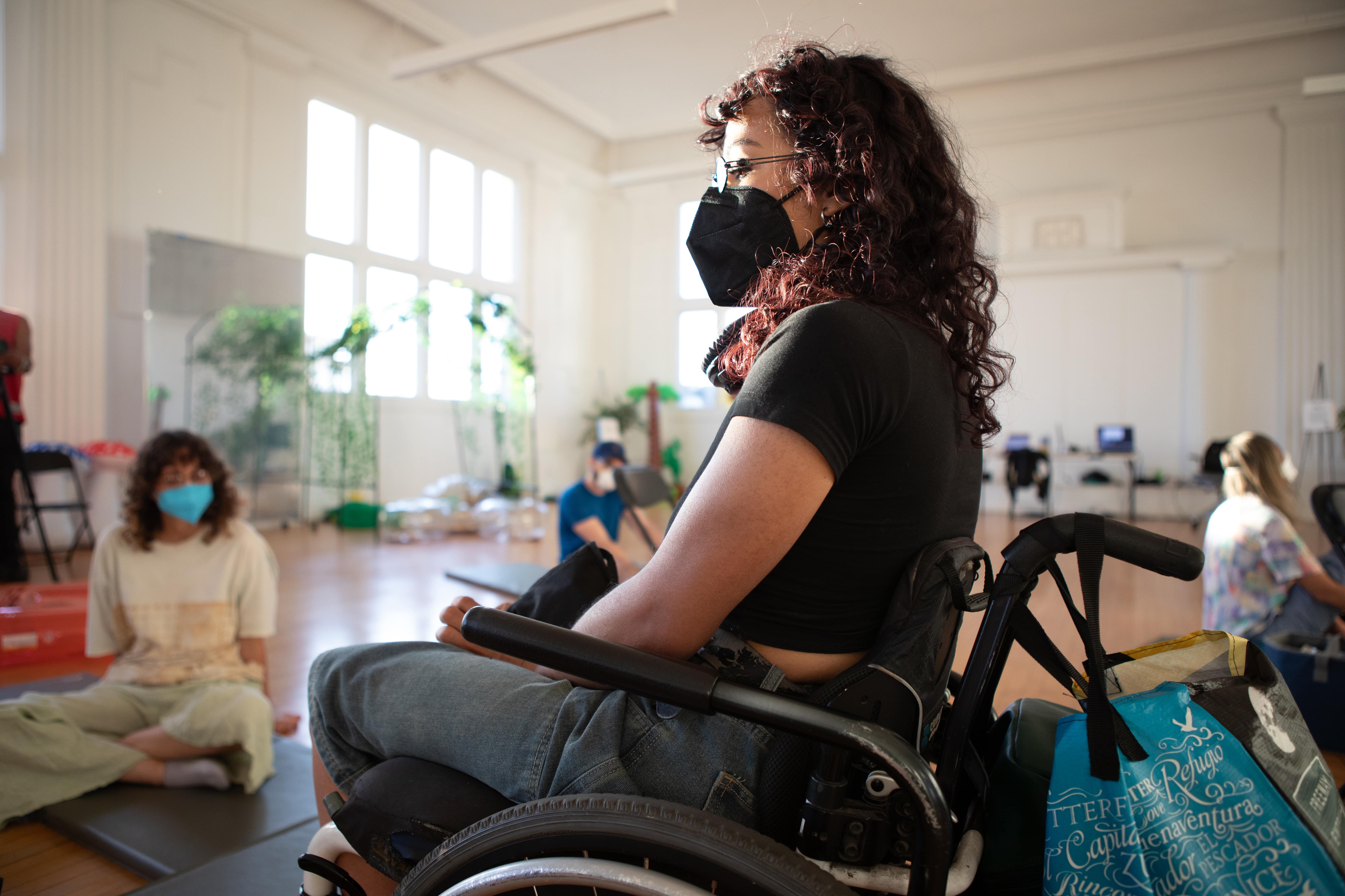 A person with dark, curly hair, wearing a black face mask and glasses, sits in a wheelchair in a sunlit room with large windows. They are looking off to the right of the frame. Another person is sitting on a mat in the background.
