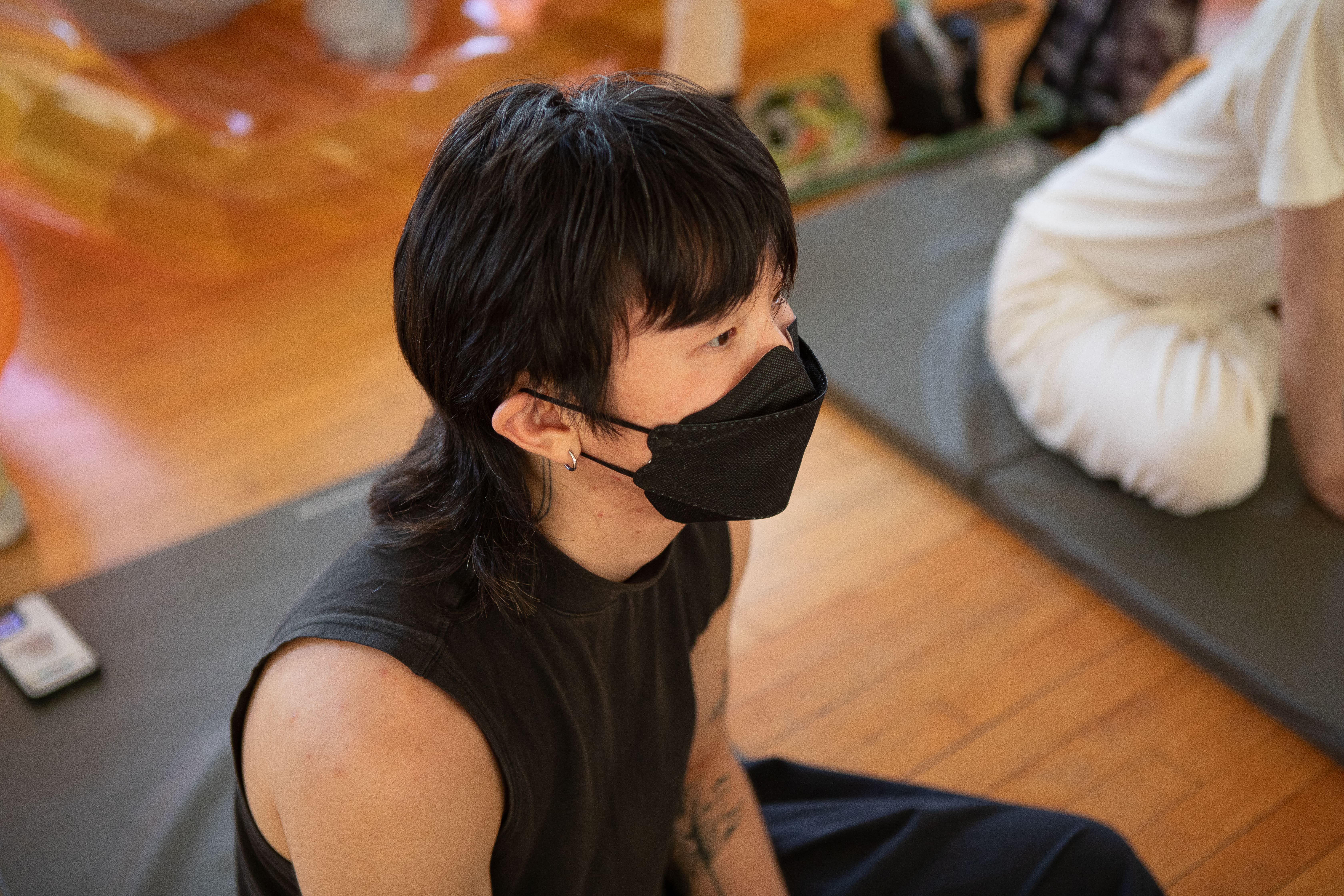 A close-up shot of a person with a mullet hairstyle, wearing a black face mask and a black sleeveless shirt. They are sitting on a wooden floor, looking to the right.