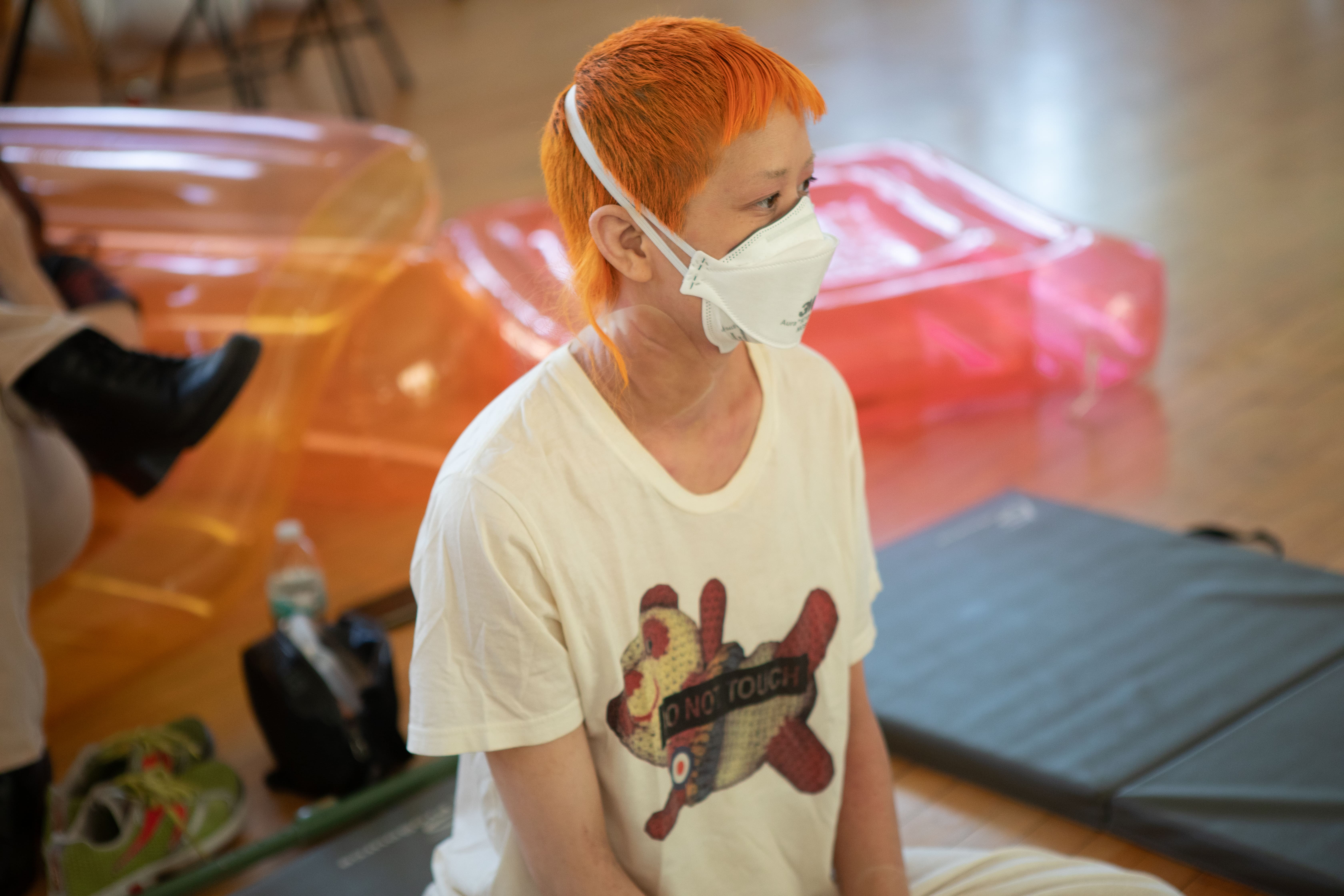 A person with short, bright orange hair wears a white respirator mask and a cream-colored t-shirt with a graphic that says "DO NOT TOUCH". They are sitting on a grey mat on a wooden floor, looking to the right.