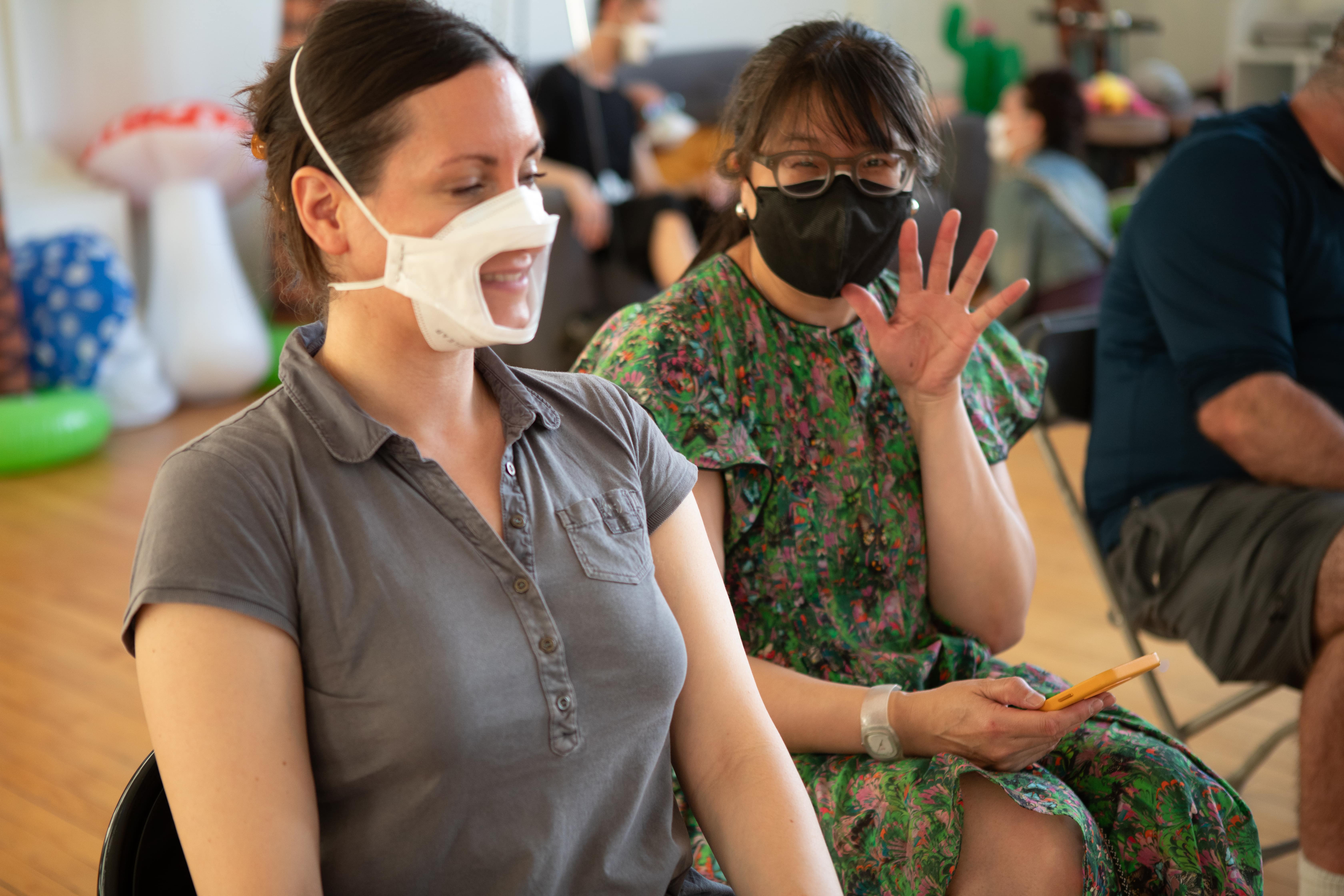 Two women are sitting next to each other. The woman on the left, wearing a grey polo shirt, is smiling and wearing a white mask that has an opening for her mouth. The woman on the right, wearing a floral dress and a black mask, is waving at the camera.
