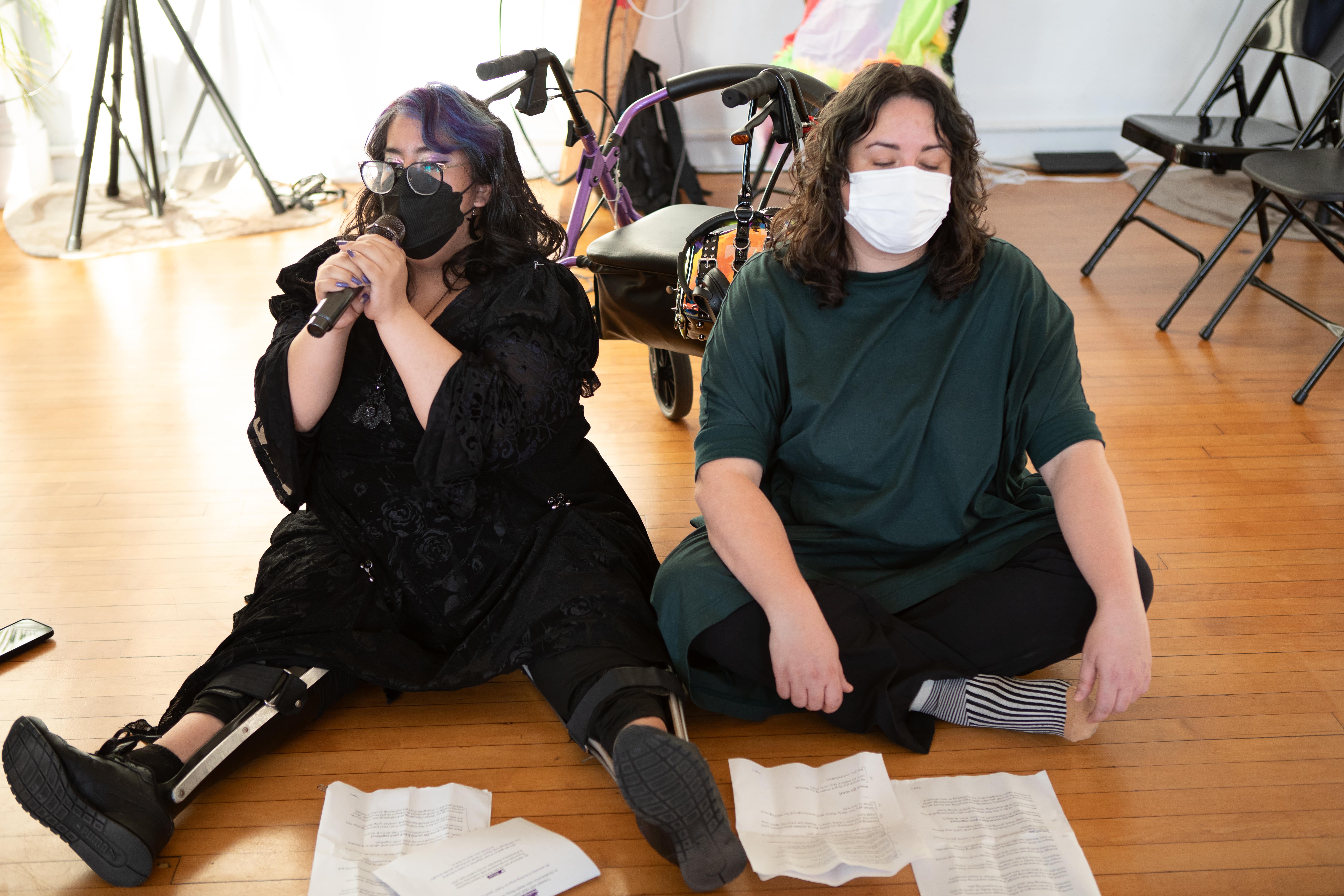 Two people are sitting on a wooden floor with papers in front of them. The person on the left, with purple-streaked hair and a prosthetic leg, speaks into a microphone. The person on the right, in a green shirt, listens with their eyes closed. A walker is behind them.