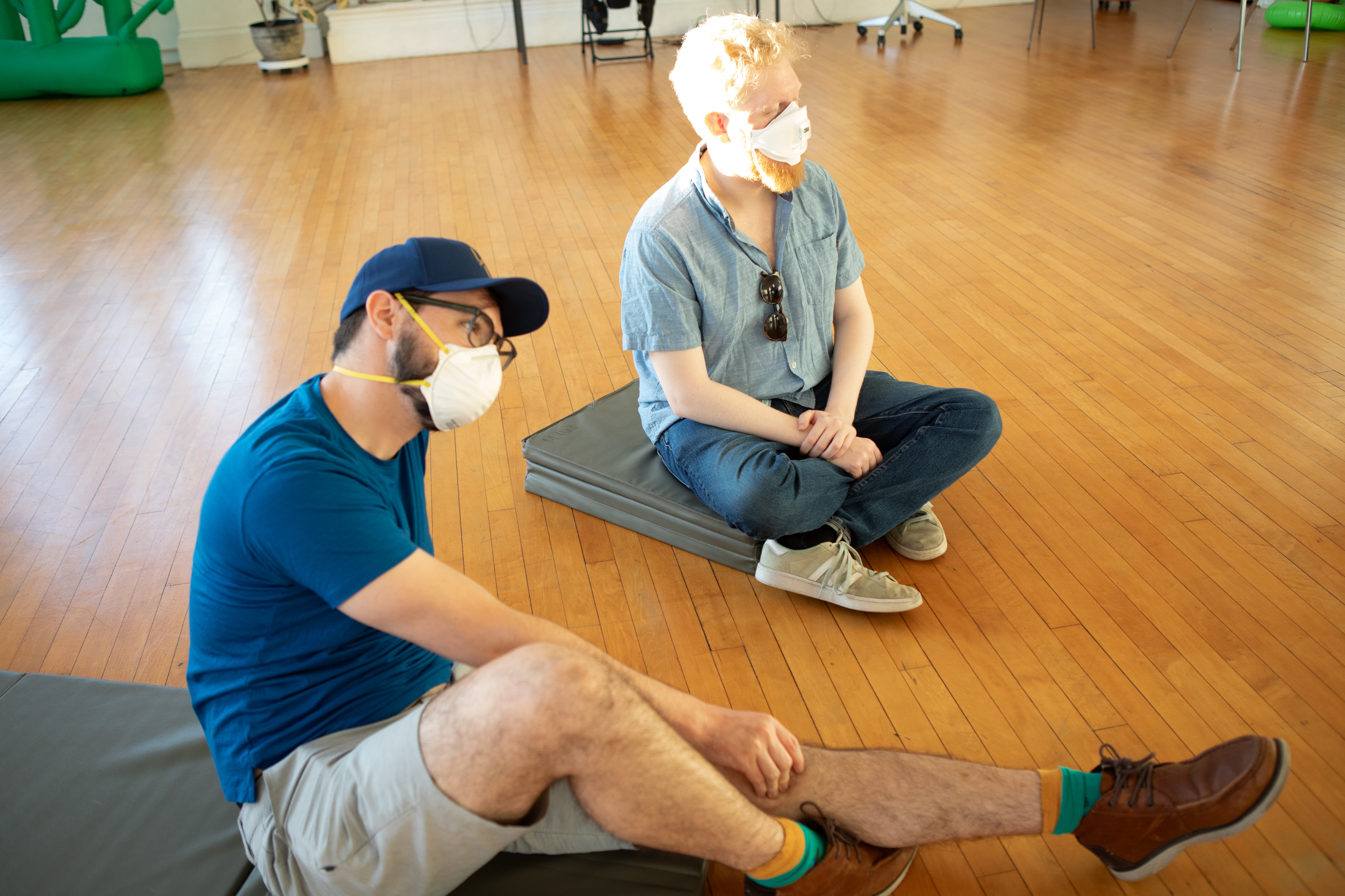 Two men wearing face masks are sitting on mats on a polished wooden floor. The man in the foreground wears a blue shirt and baseball cap, while the man behind him has blond hair and wears a light blue button-up shirt.