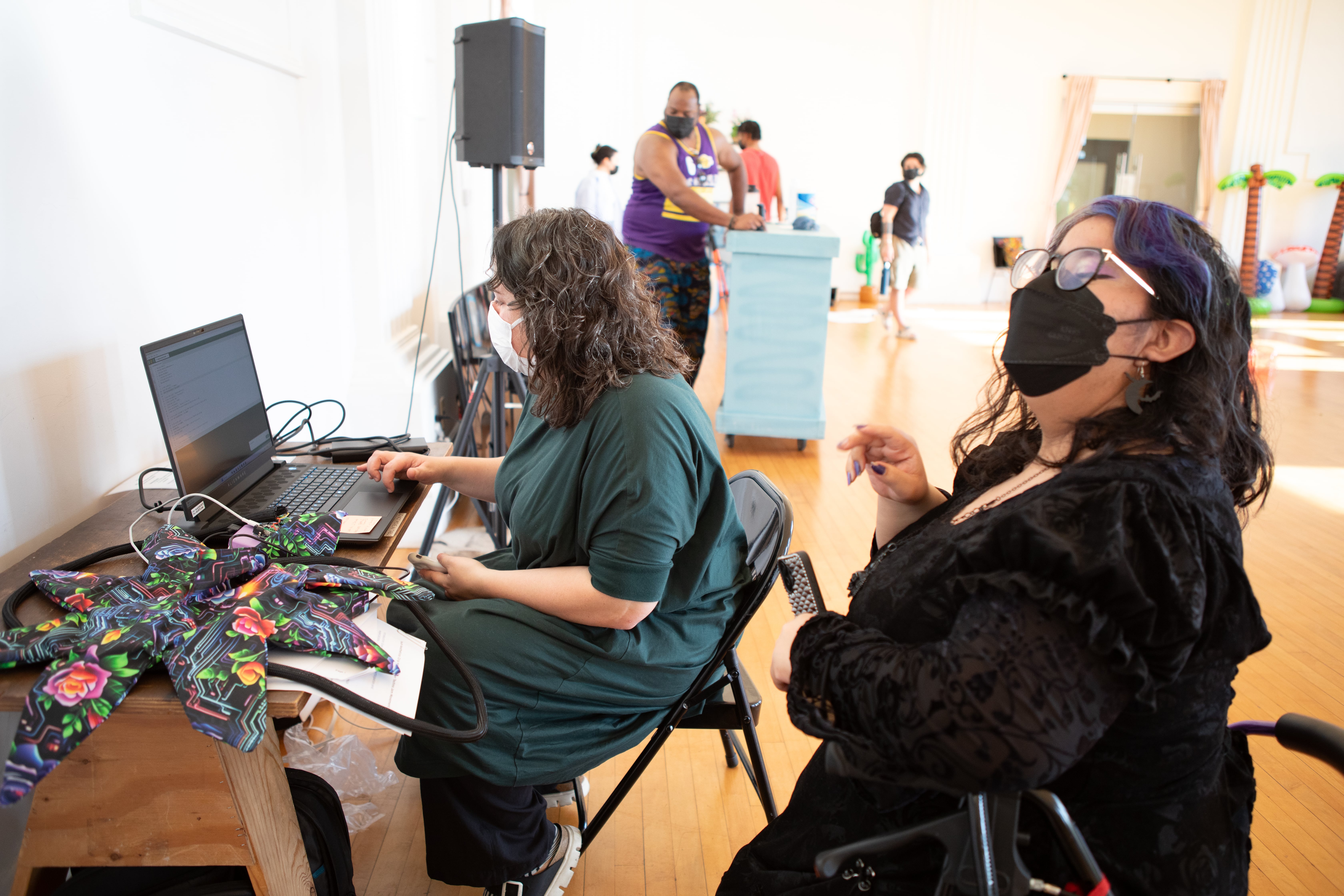 Two women wearing face masks are at a desk with a laptop. The woman on the left, with curly brown hair, is using the laptop. The woman on the right, with purple-streaked hair and glasses, is in a wheelchair and looks toward the right side of the frame. In the background, other people are in a large, open room.