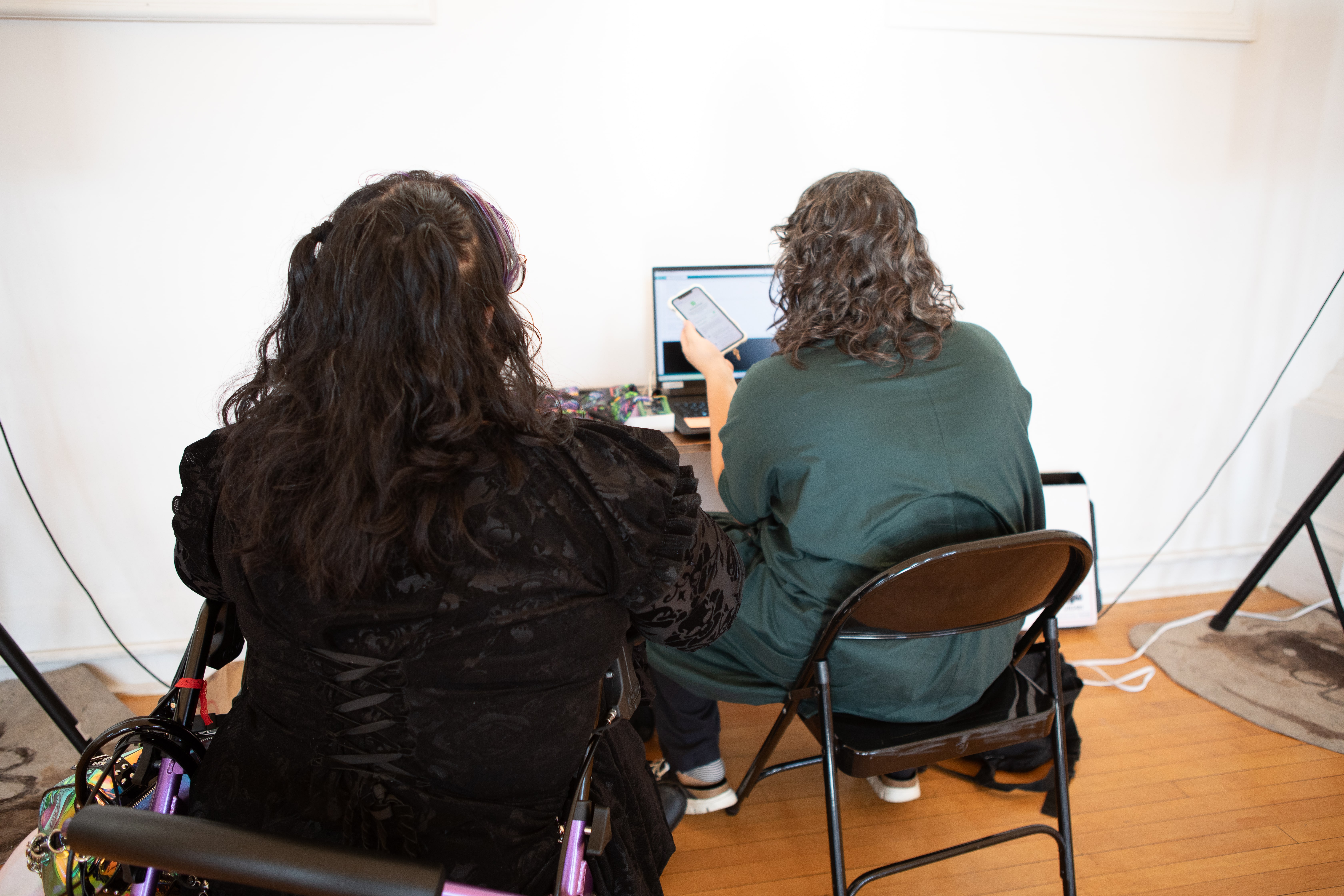 The back of two women sitting at a desk and looking at a laptop screen. The woman on the left has long, dark hair with purple highlights and is in a wheelchair with a walker behind her. The woman on the right has long, curly brown hair and is showing something on a phone.