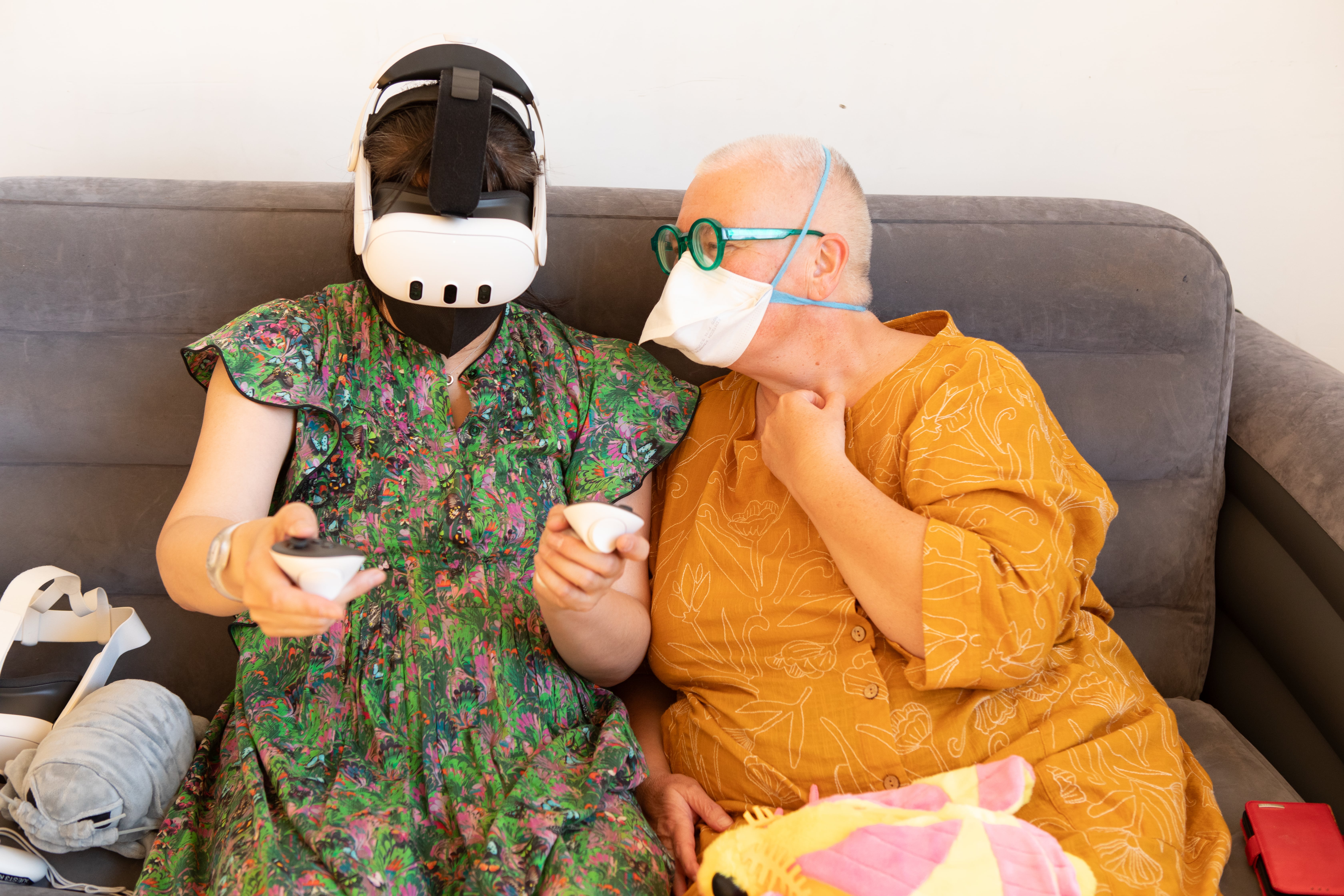 Two people are sitting on a grey couch. The person on the left is wearing a VR headset and a colorful floral dress and is holding controllers. The person on the right, wearing a yellow top and a face mask, is looking at them with a hand to their chest.