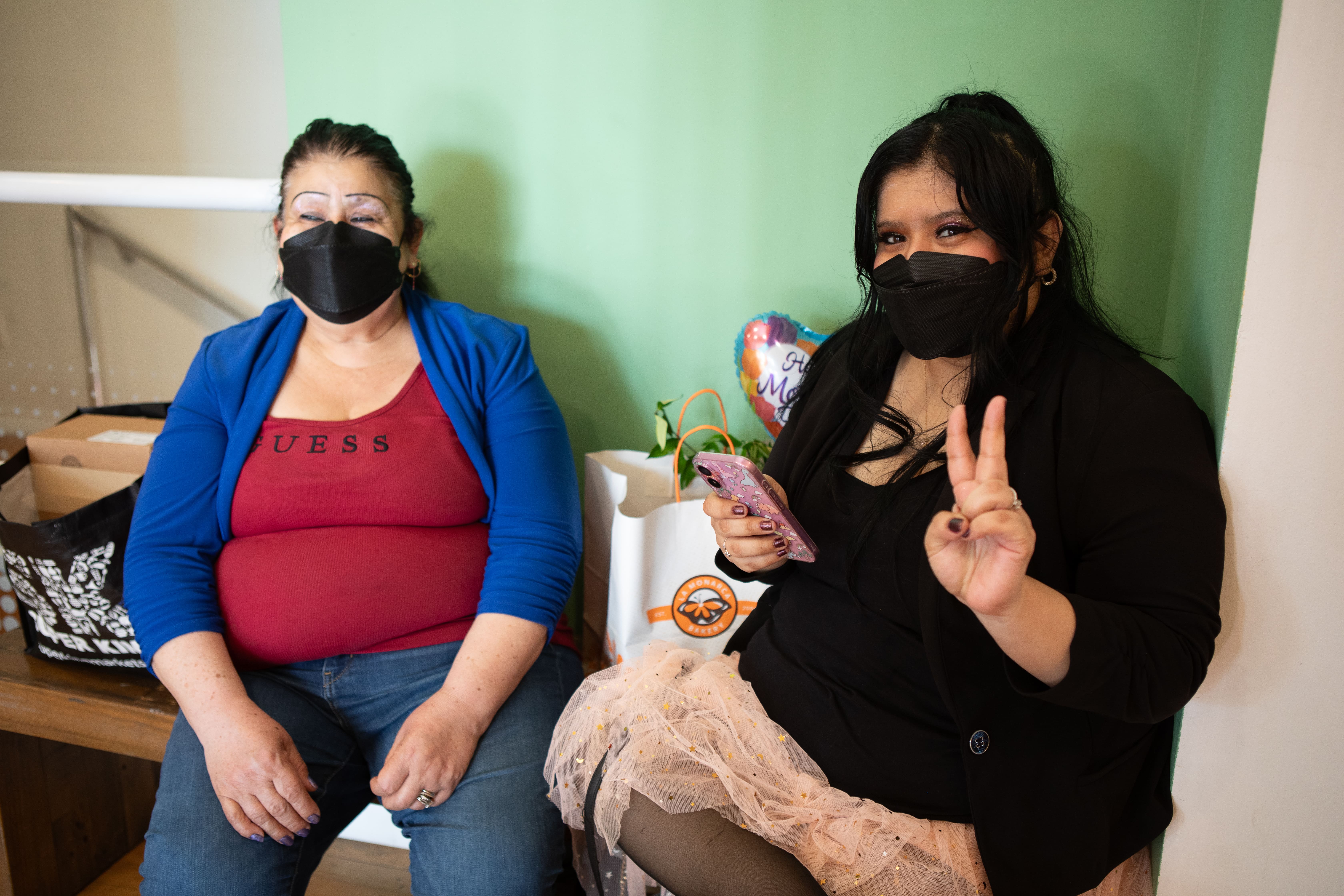 Two women wearing black face masks are sitting next to each other against a light green wall. The woman on the left wears a red shirt and a blue cardigan. The woman on the right, wearing a black top and a pink sparkly skirt, holds up a peace sign and a cell phone.