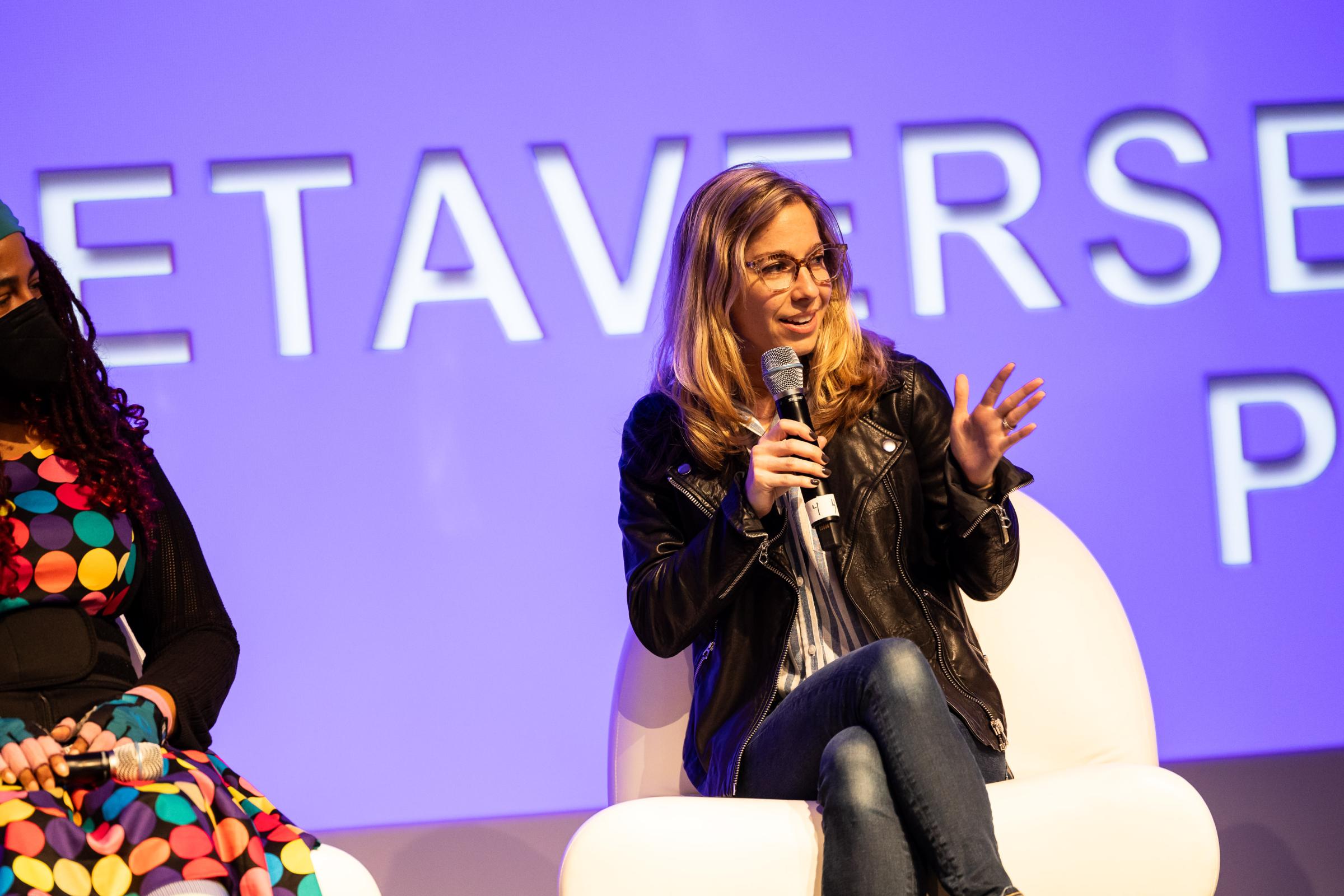 Female presenting individual with glasses and blonde hair wearing black leather jacket sitting on a white couch, talking while holding a microphone. 