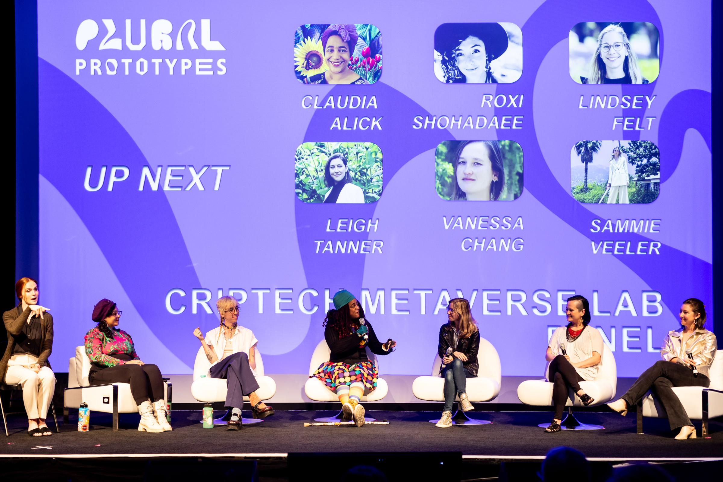 Panel of 7 individuals, on the left is an ASL interpreter seated. Behind the panelists is a giant purple screen with their names, profile pictures, and event name. 