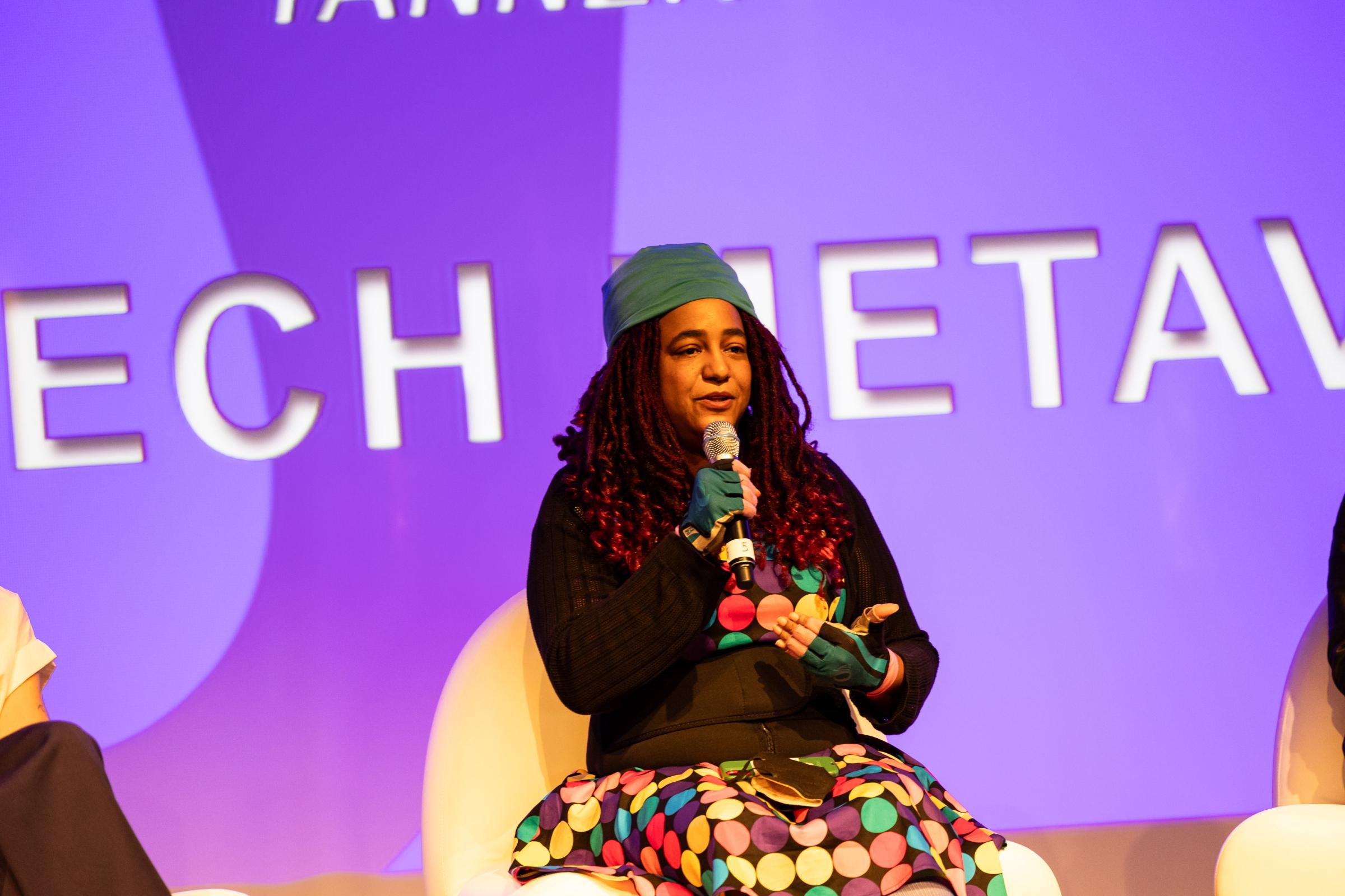 Female presenting individual with dark red hair wearing a green cap and a multicolored dress. Sitting on a white couch speaking while presenting and holding a microphone. 