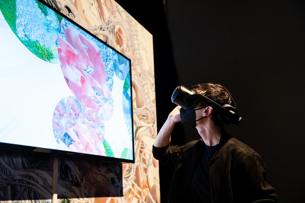 Individual with pale skin and brown hair is wearing a dark brown tshirt and black mask. They are wearing a VR headset facing a screen showcasing orbs with coinciding colors of green pink and blues. On the back is a similar wallpaper but with beige and white tones.