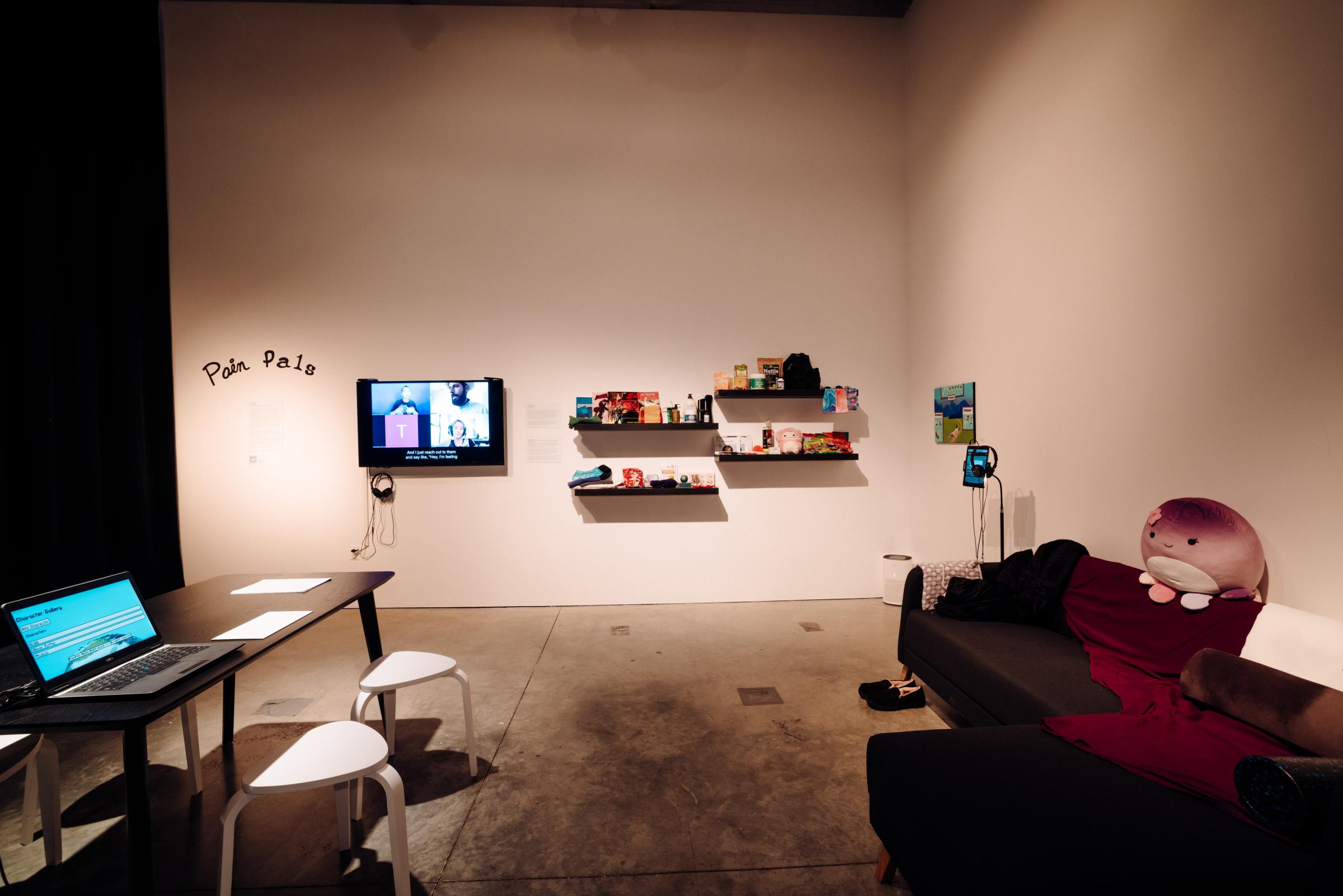 A dining table with four low white stools is positioned in front of a large wall-mounted screen.Floating black shelves with an assortment of self-care items bear a variety of items, including stuffed animals, snacks, pamphlets, heating pads, lotion, compression socks, Vitamin C packets, comic books. Against the gallery’s back wall is an L-shaped upholstered dark gray couch and a cozy robe. An iPad is mounted on a stand with headphones.

