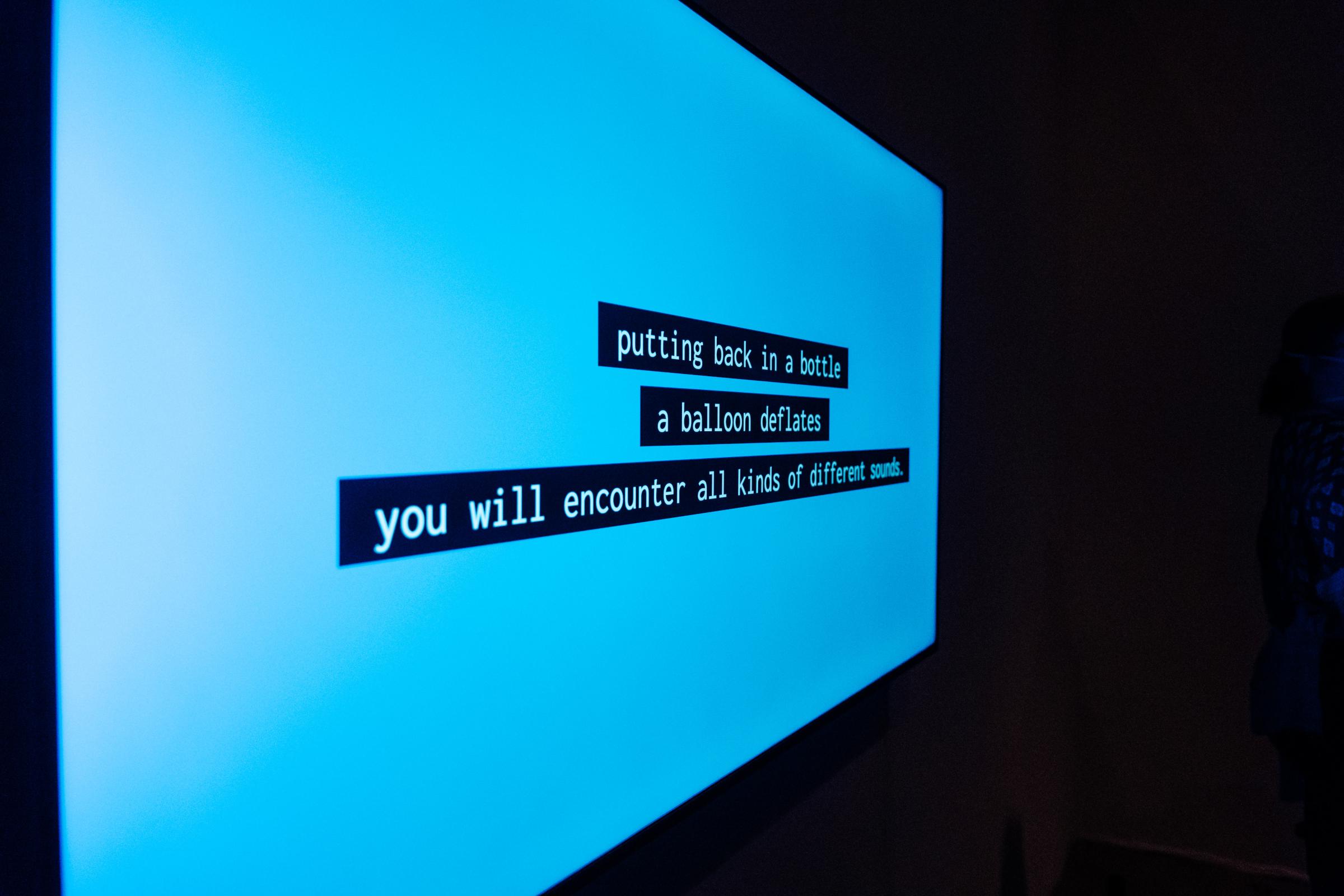 Diagonal shot of a monitor with a blue screen. White text with black highlight displays in the center reading "putting back in a bottle a balloon deflates you will encounter all kinds of different sounds."