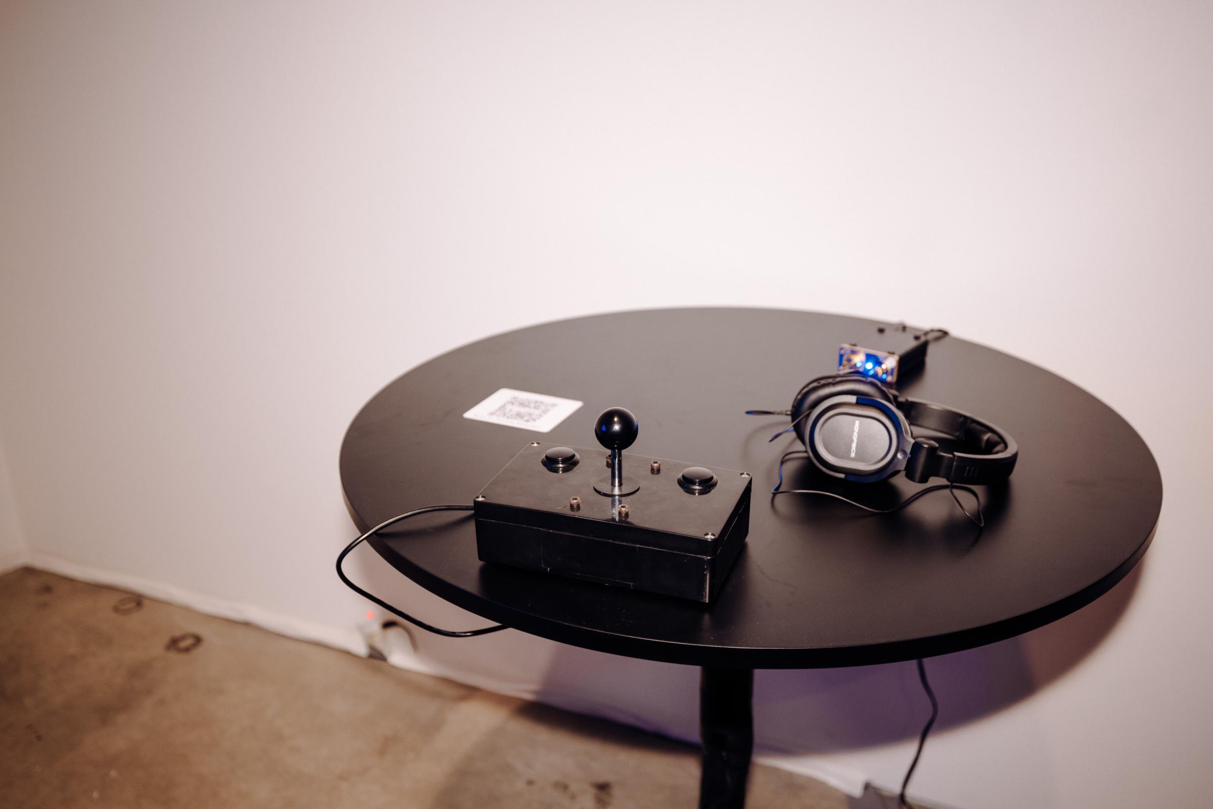 Circular table with joystick and buttons on a controller. There is a headphone amp with lights on and a pair of headphones directly in front of the controller. To the left of the controller is a QR code. 