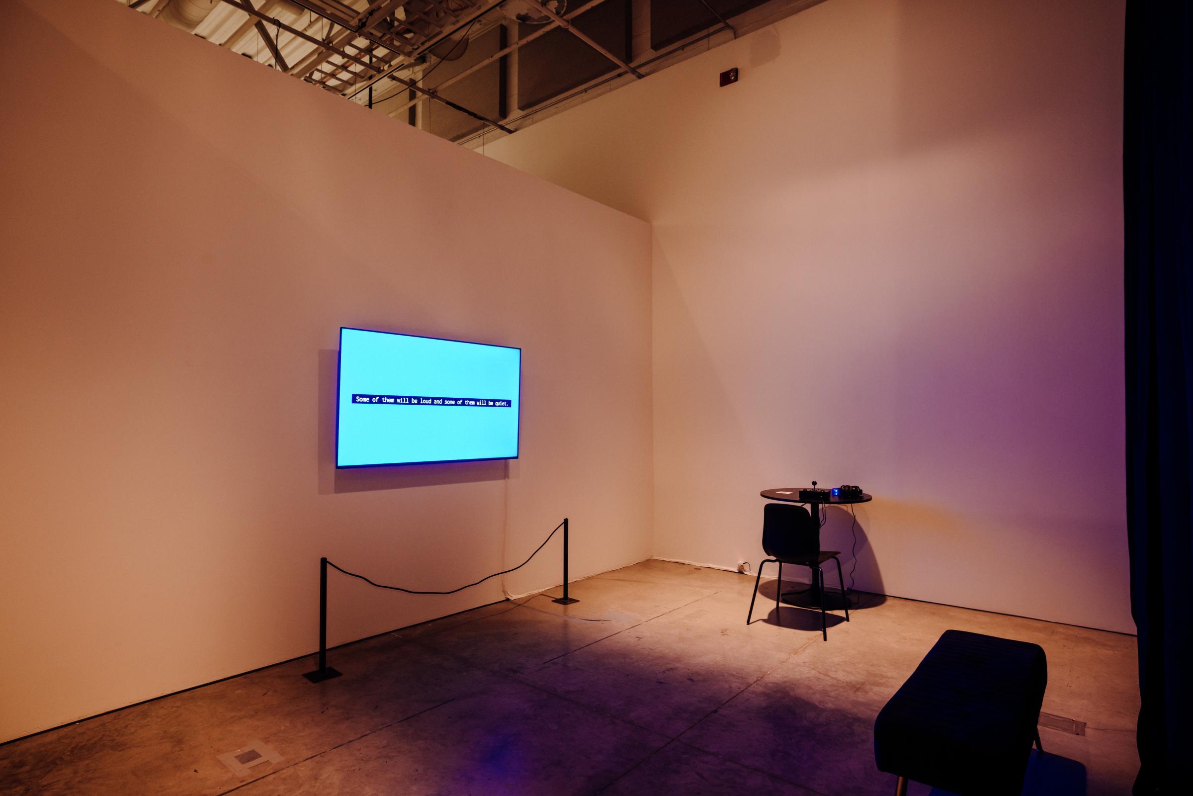 Room with a circular table against the back wall. A chair sits in front of the table, facing the wall. A large monitor displays text “Welcome to Unseen Sound.” Across from the monitor is a short bench. 

