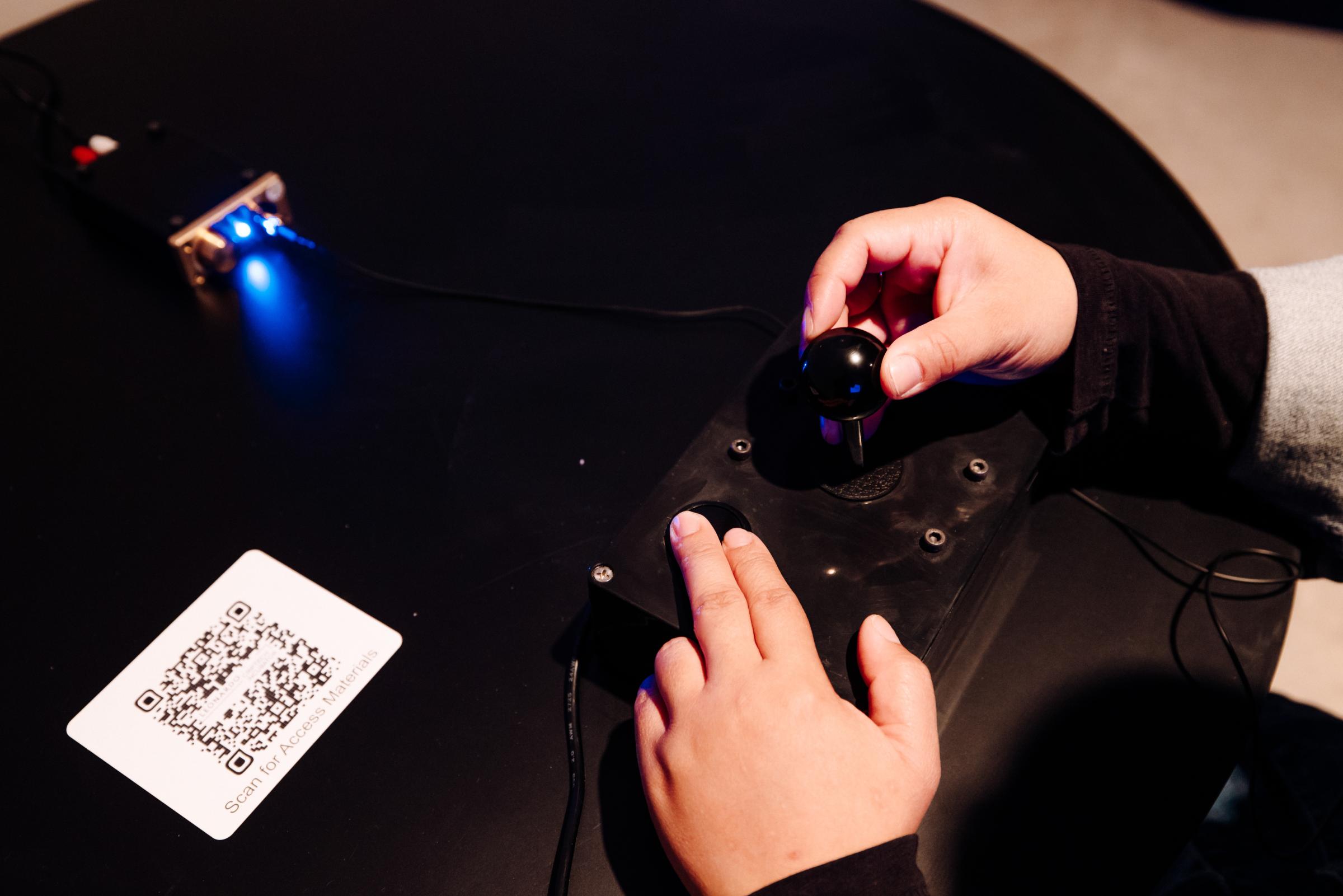 Visitor with pale hands is interacting with a joystick and buttons on a controller. There is a headphone amp with lights on directly in front of the controller. To the left of the controller is a QR code. 

