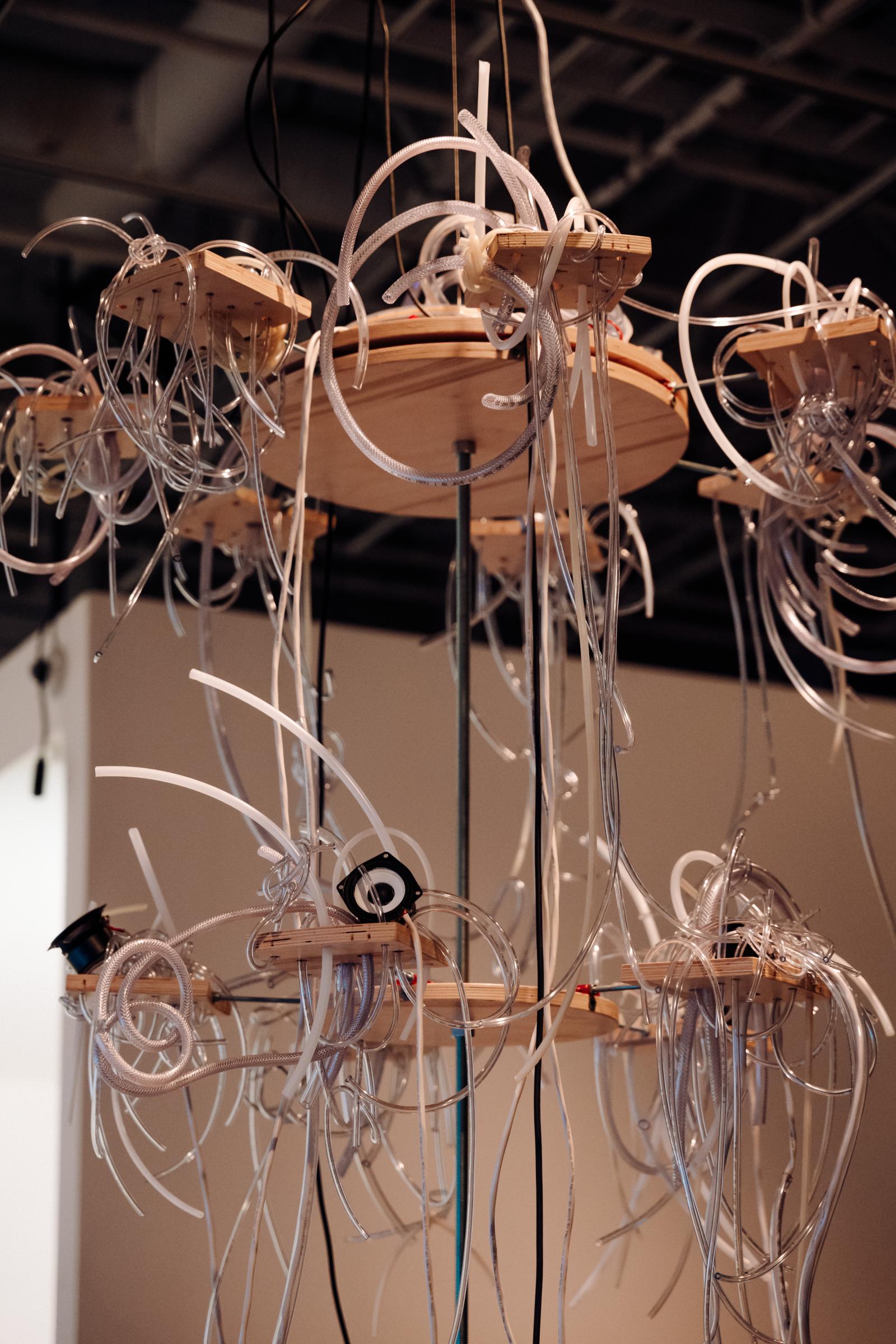 Multi-tiered suspended wooden chandelier with plastic tubes spilling outwards over the sides. There are four speakers and two microphones integrated into the sculpture. 

