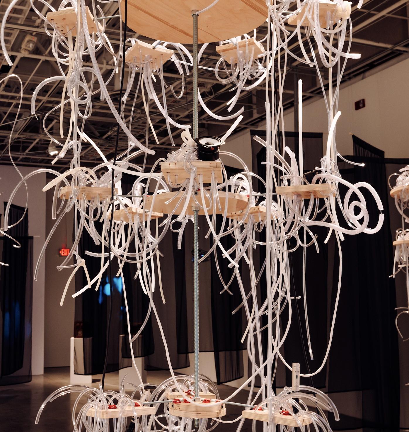 Close-up of babbel’s central chandelier, a multi-tiered suspended wooden sculpture with plastic tubes spilling outwards over the sides. There are four speakers, two microphones, and red 3D printed parts integrated into the sculpture.