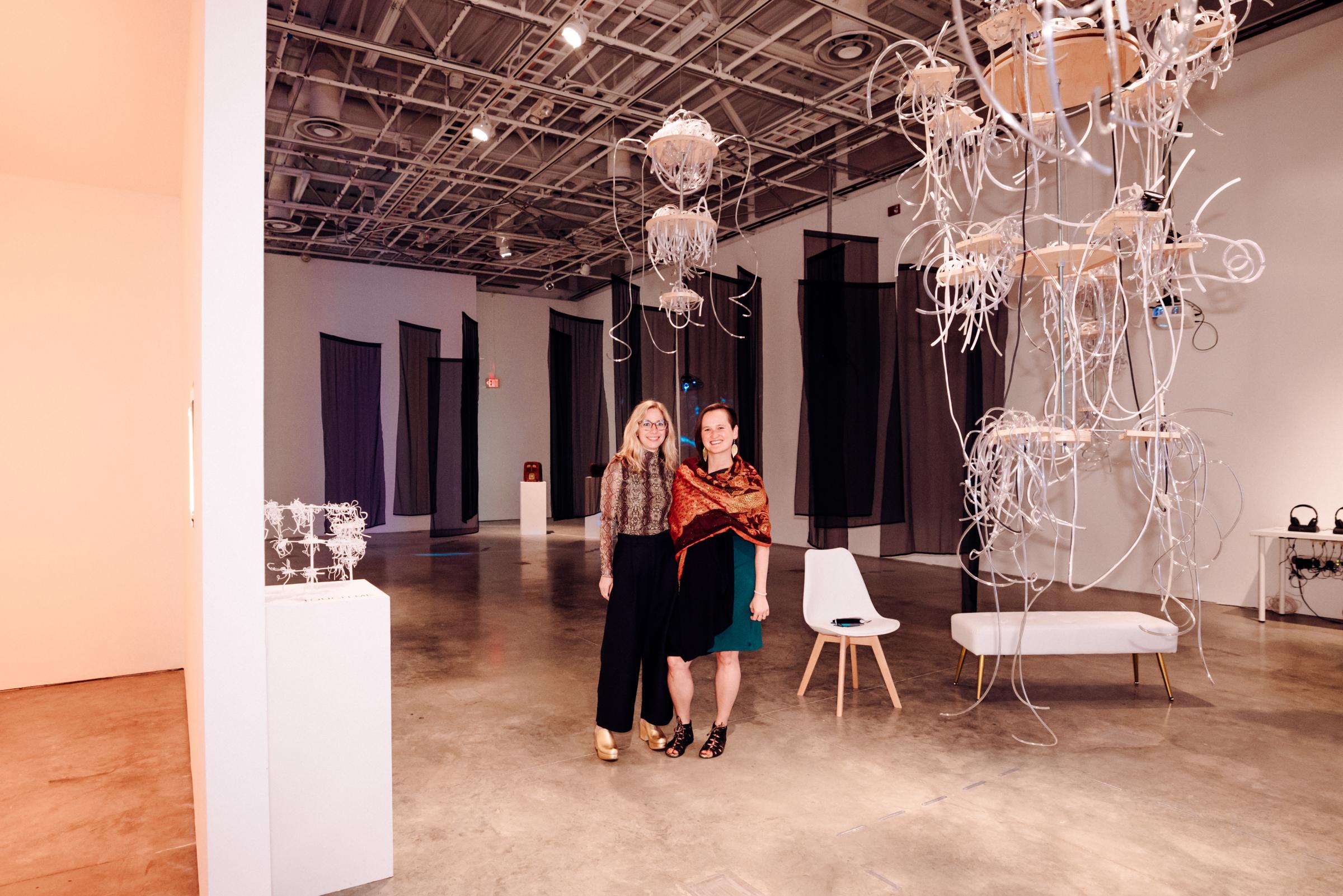 A blonde haired woman and a brown haired woman stand together in the middle of the E.A.A.T. gallery space. 

