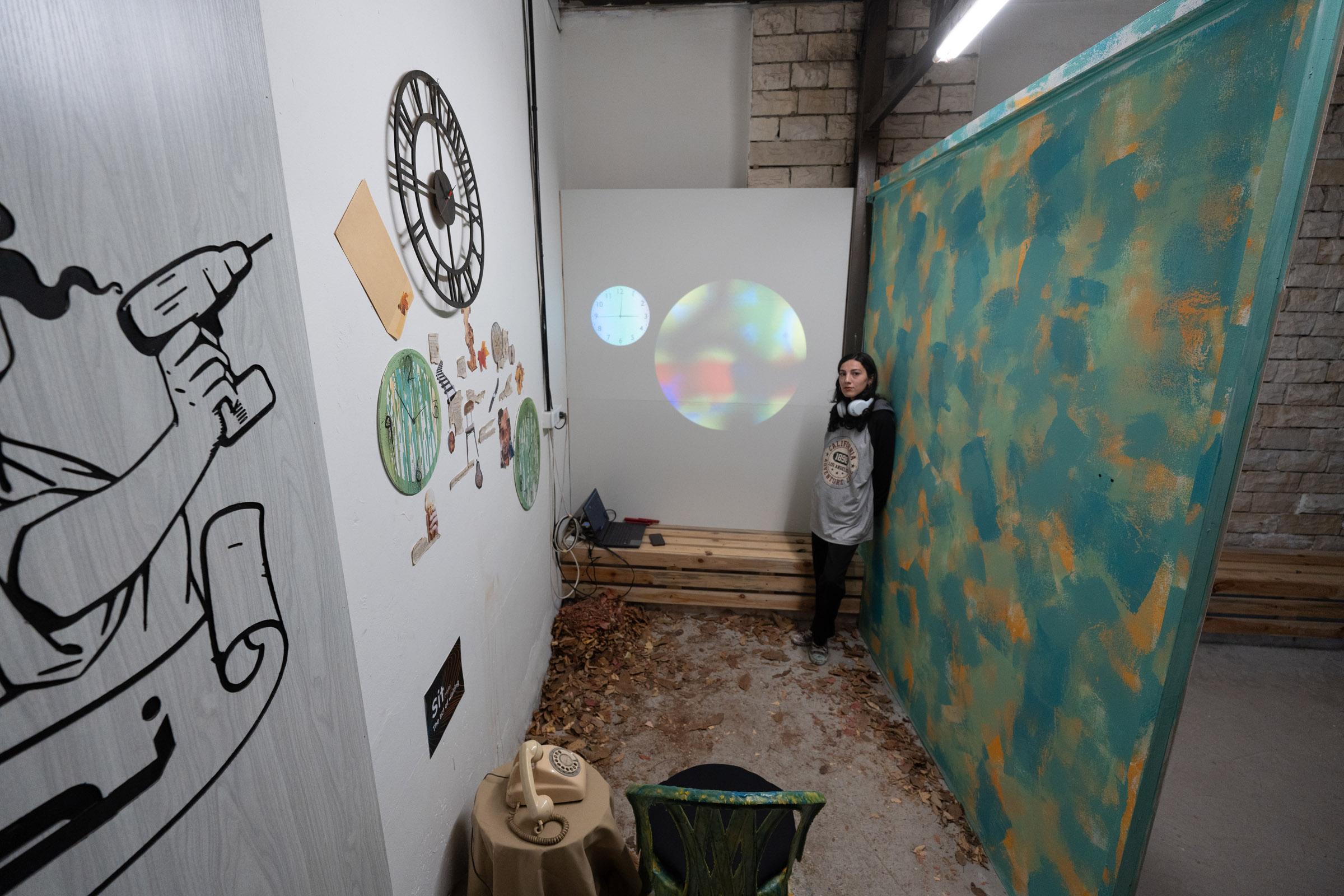 Room view from above showing a wall with various circular and other mixed media elements, a central projection, and a multicolored wall. In the back corner is an individual wearing headphones and a white colored garment.