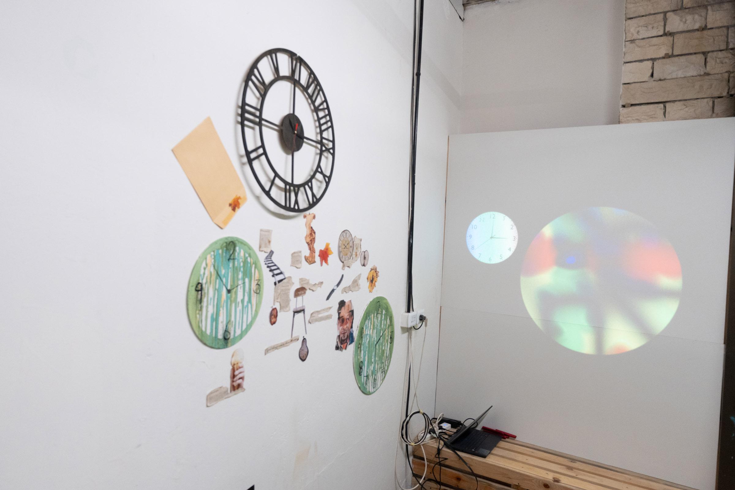 Wall with various items attached with three big clocks the most prominent. Interspersed amongst are mixed media collages depicting cutouts of different items, including leaves, railroad tracks, and chairs. Adjacent is a large wall with a small clock projected and a larger circular projection with black, yellow, red, and green hues.