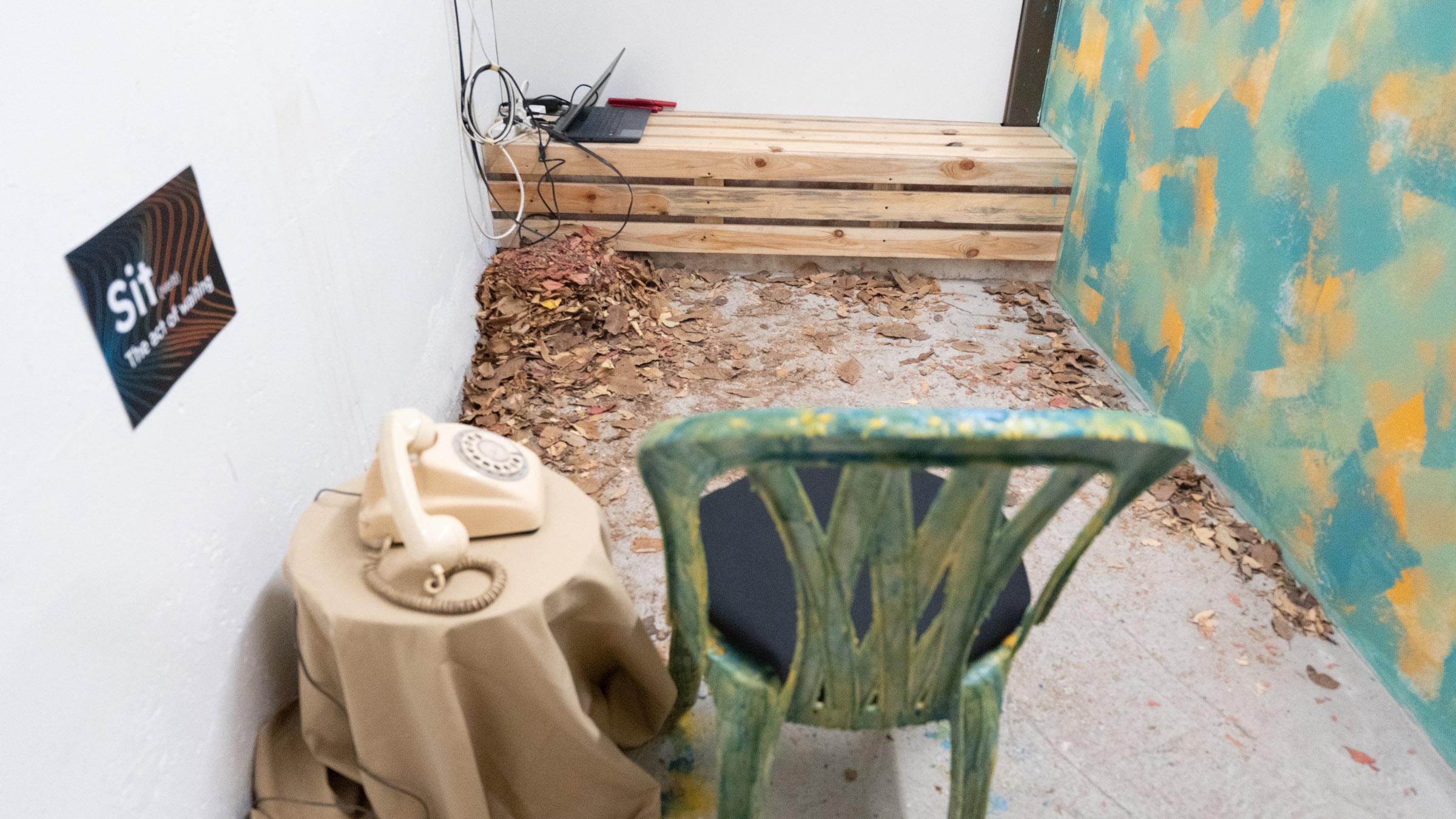 A plastic chair with pastel hues of blue, green, and orange parallel is a wall painted in the same manner. A side table accompanies the chair with a white rotary telephone, and a black sign above reads “sit: the act of waiting,” across from the chair is a wooden bench with leafy foliage under it.