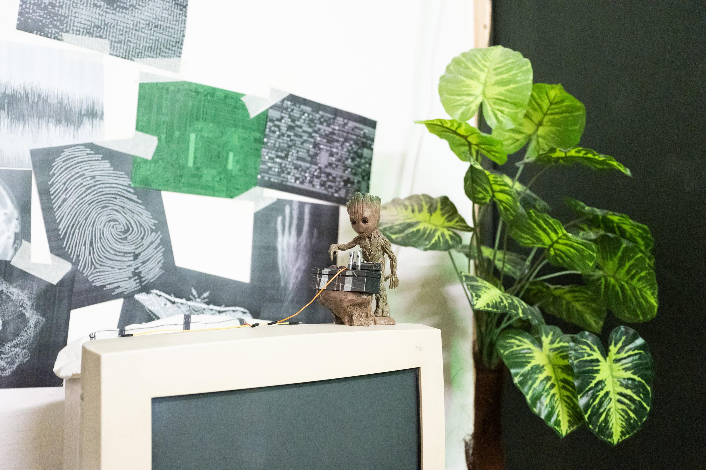 Close-up of an action figure on top of a computer monitor interacting with a miniature set. In the background is a large plant.