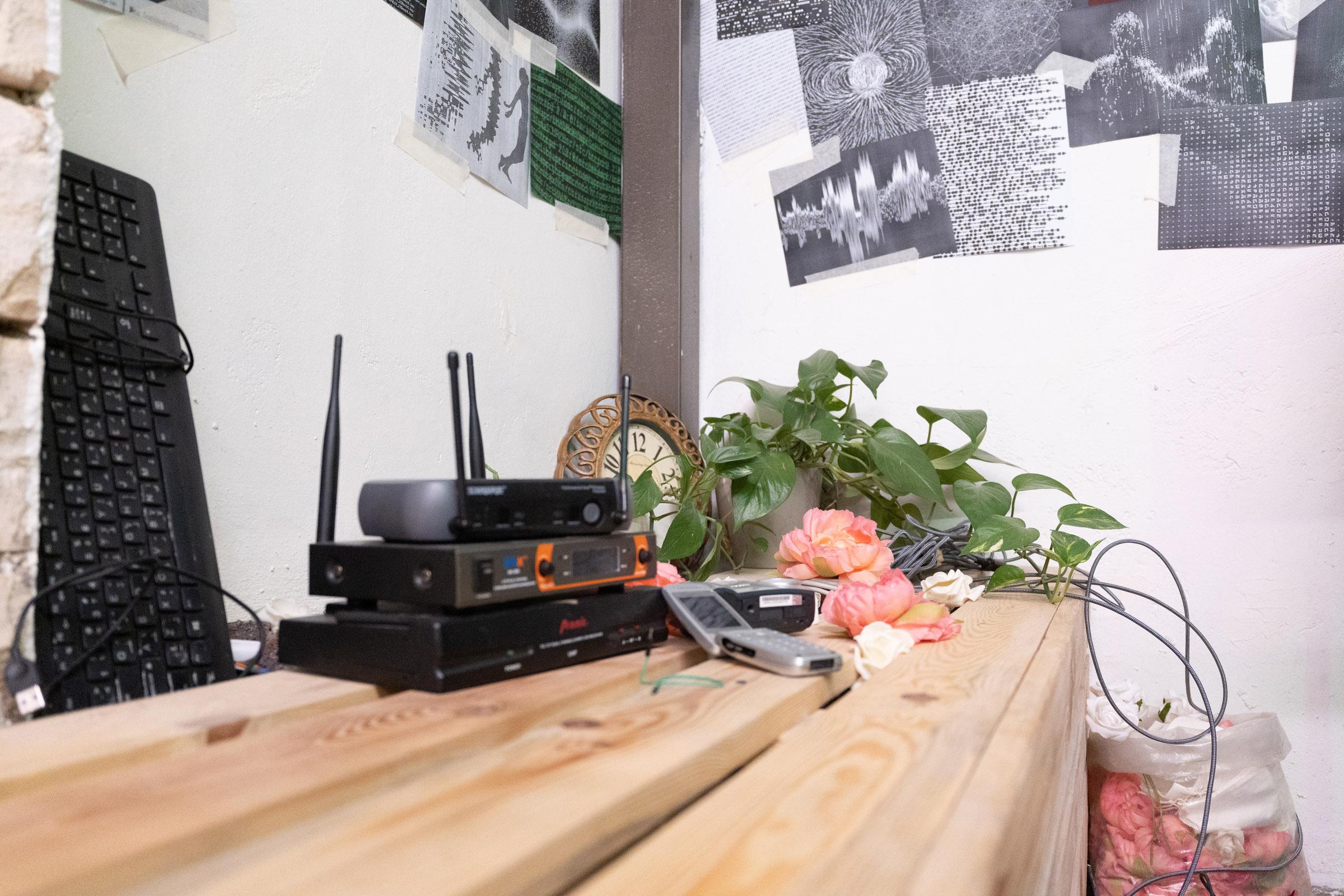 Close-up of wooden bench with three modems and radios in the center, surrounded is a keyboard and plants. In the background is a clock and posters on a wall.