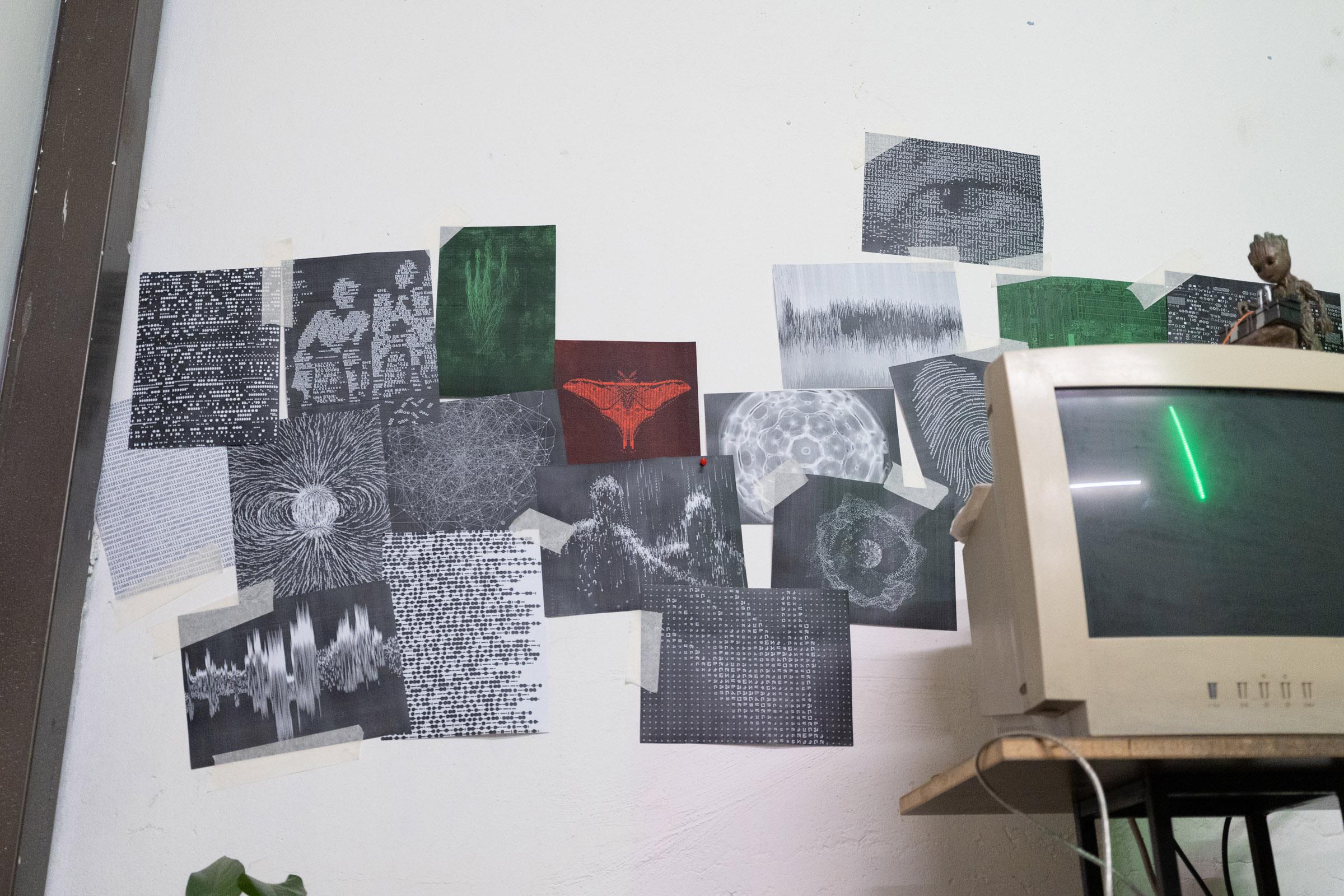 Various cymatic images in grey, green, and red are plastered on the wall, on the right is an old computer monitor with a single short green line running vertical.