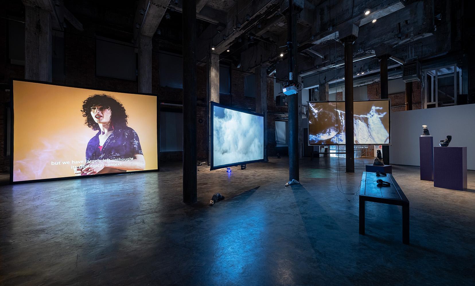 Wide gallery view, main view is a screen projecting someone with pale skin, black curly hair, and a purple shirt. Another screen shows white clouds, interspersed are various art items. 
