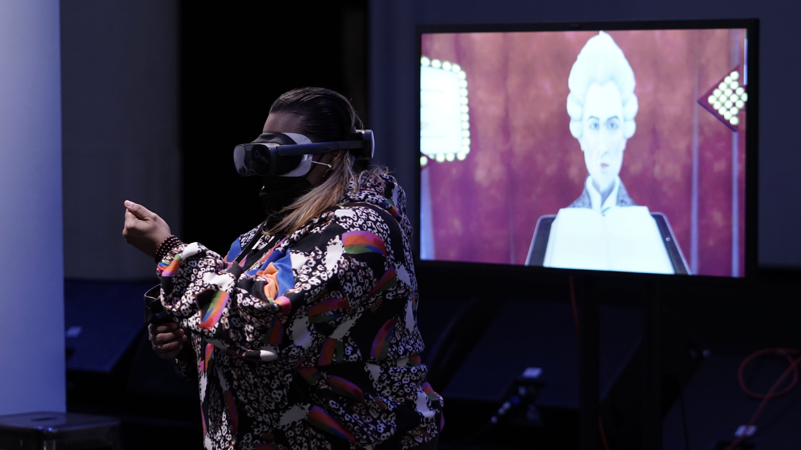 Graphic designer and disability justice advocate Jen White Johnson, an Afro-Latina woman in a colorful jacket wearing a VR headset, stands in front of a screen that shows her perspective in the virtual world of Maestro. The screen shows a white man in a wig standing behind a conductor’s lectern
