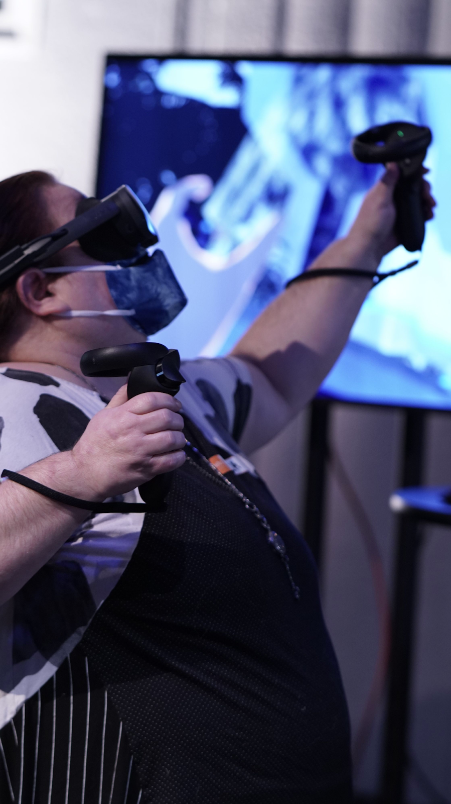 a white woman wearing a black-and-white dress, stands in front of a screen that shows her perspective in a virtual world. Scott is wearing a VR headset and carries one controller in each of her hands, one of which is held above her head as if pointing to something
