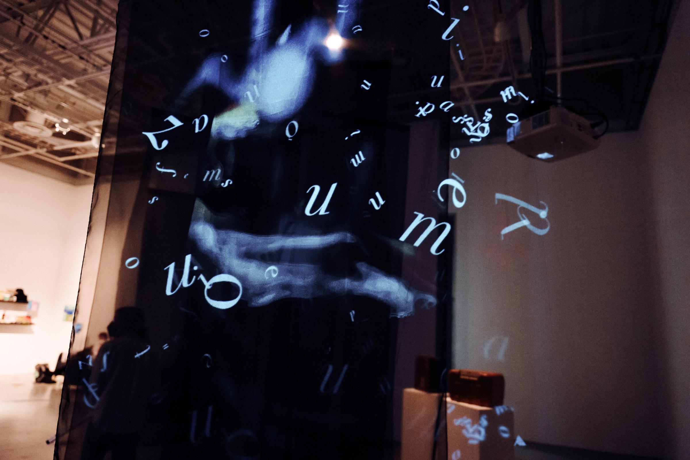 Projected in white onto translucent black fabric, a spray of letters pattern the blurry image of a piano player. 

