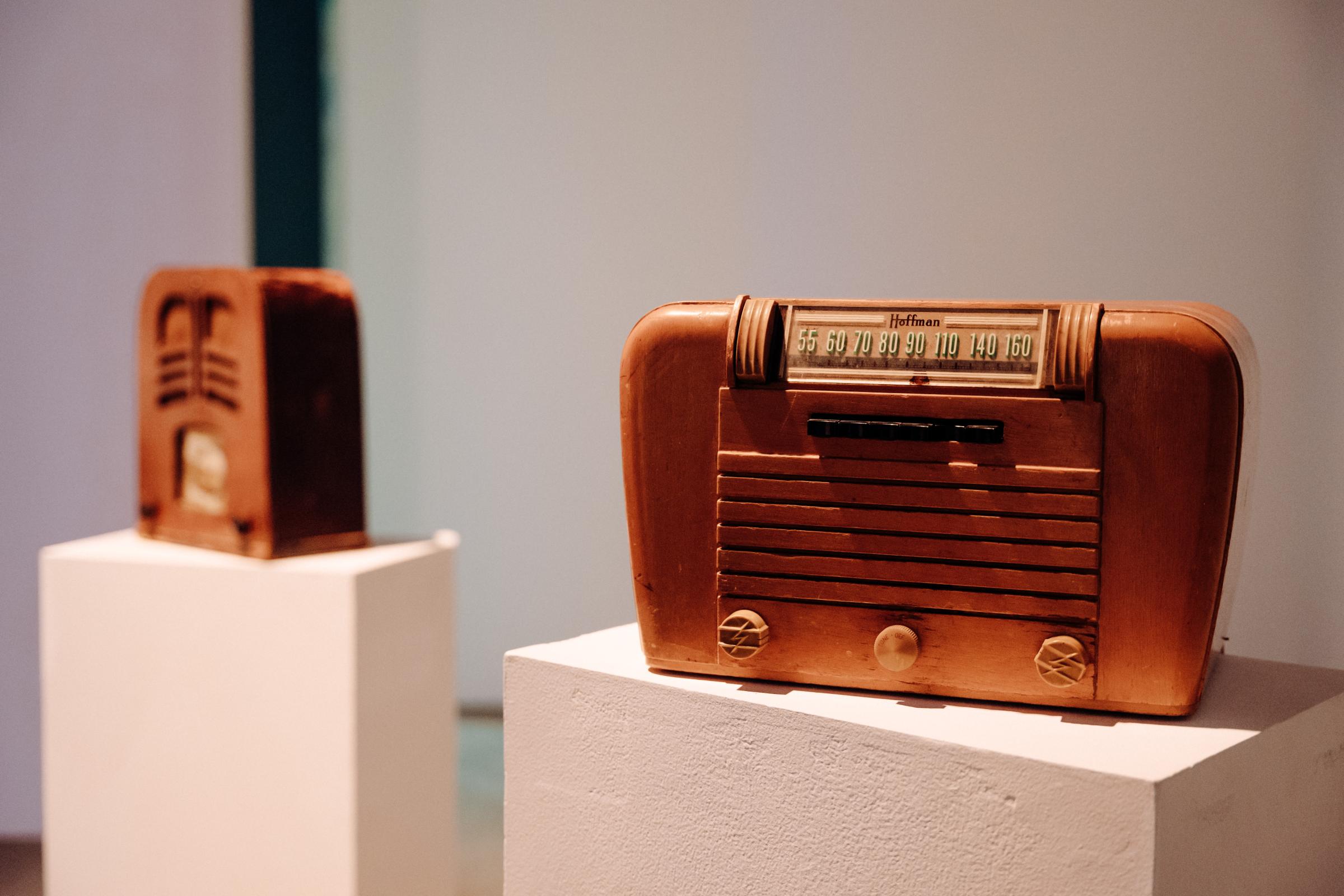 Two wooden vintage radios on white pedestals. The one in the background stands vertical, the one in the foreground is wide with three knobs. 


