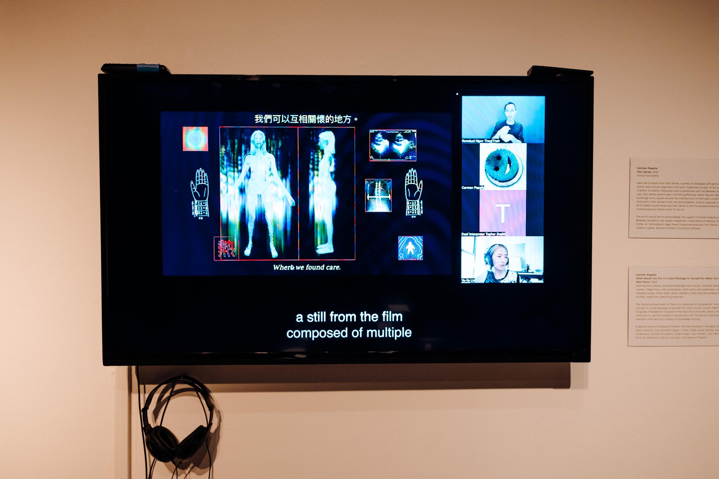 Monitor in the center showcasing an X-ray image of a female body on a zoom call. At the bottom of the monitor hangs a pair of headphones that connect to the monitor. To the right are two wall text blurbs. 

