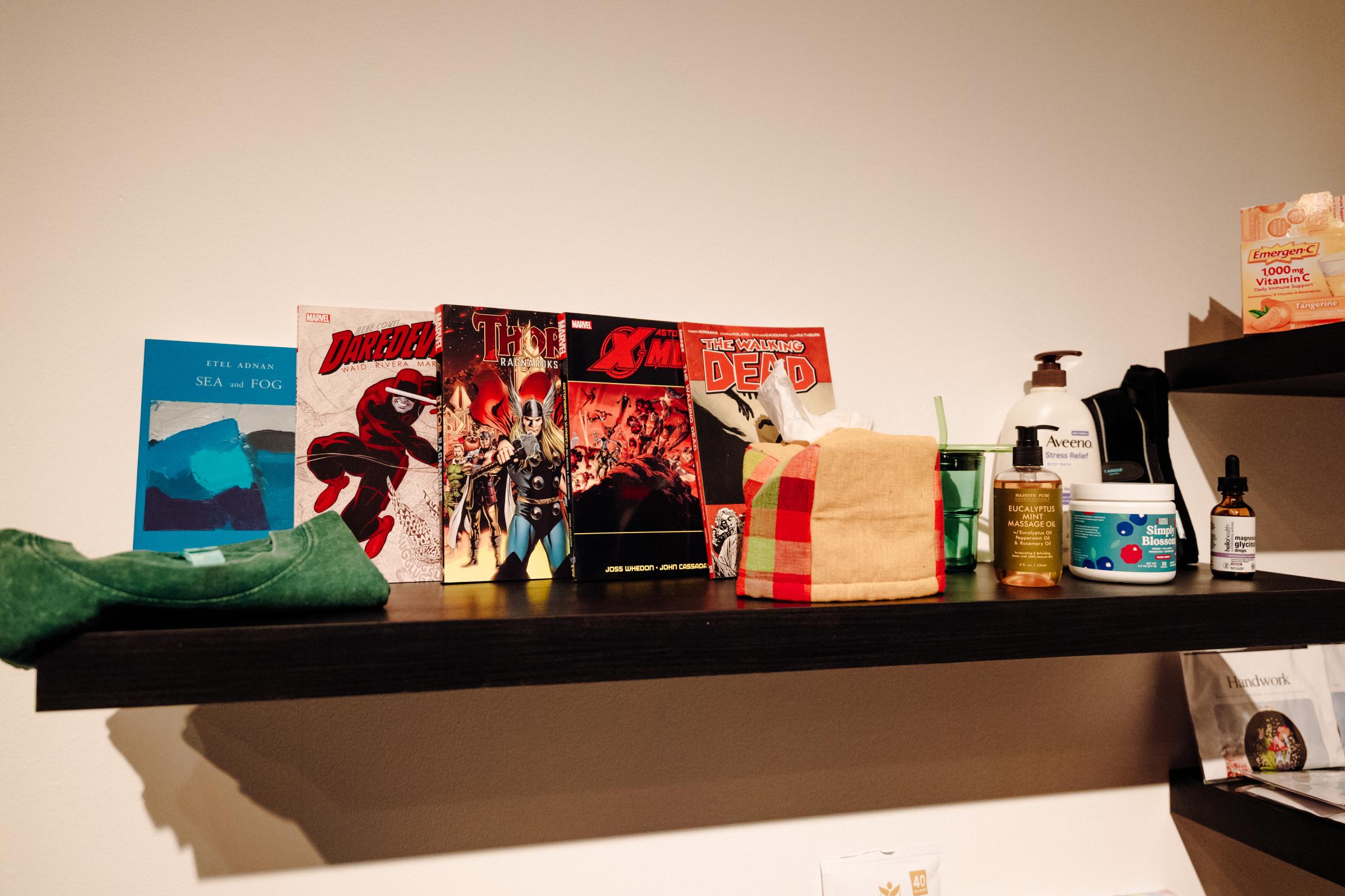 Shelf with various items including, comic books, folded green tshirt, checkered tissue box, cup, lotions, tinctures, amongst others. 