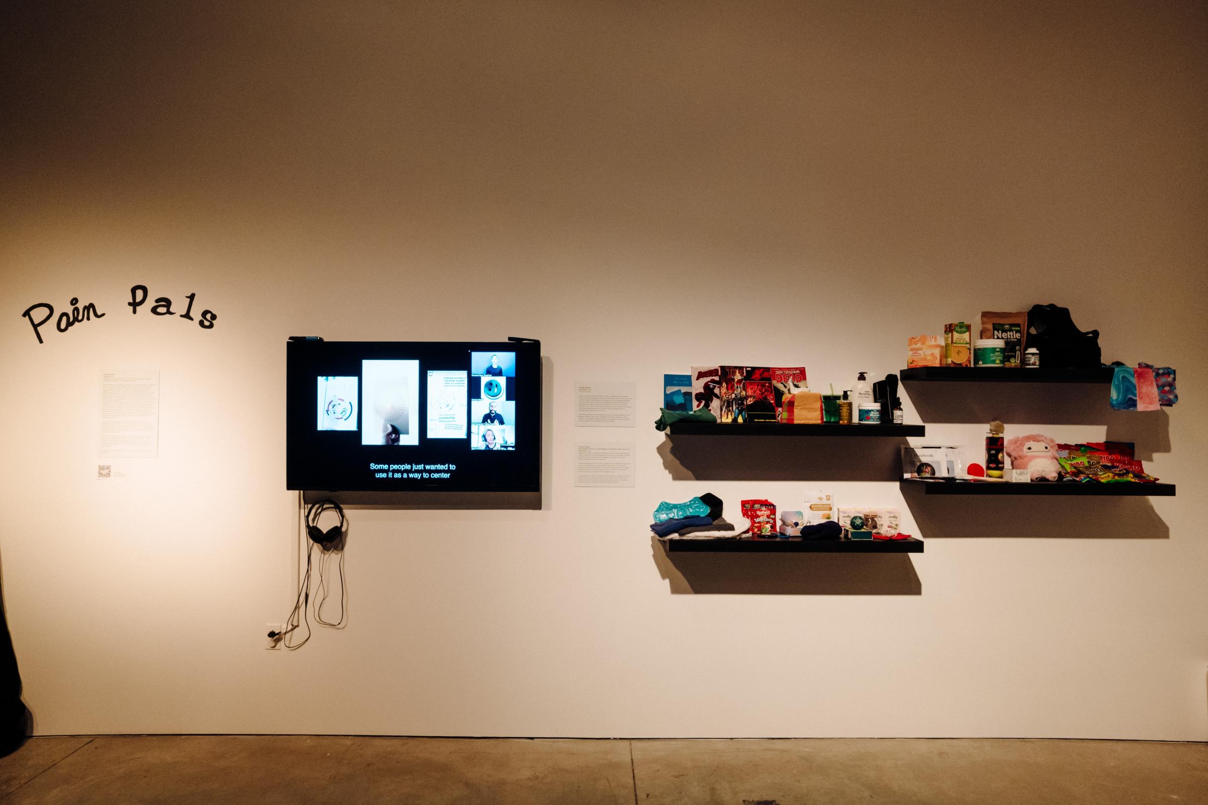 Floating black shelves with an assortment of self-care items bear a variety of items, including stuffed animals, snacks, pamphlets, heating pads, lotion, compression socks, Vitamin C packets, comic books. A monitor displaying a zoom call with someone sharing a screen; underneath is a pair of headphones. 
