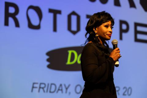 Individual wearing with black hair and black dress is standing on stage holding a microphone. Behind is a blue banner screen. 