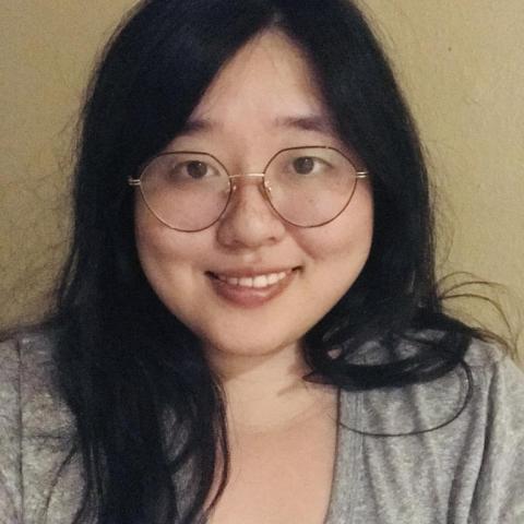 Iris, a light skinned Asian person with long, wavy black hair and gold and black rimmed hexagonal glasses, is smiling directly at the camera. They are wearing a deep v neck light-medium gray t-shirt