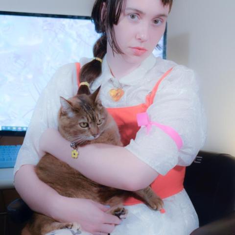 Nat, a white non-binary person with dark brown long hair and step bangs, sits on an electric scooter holding their reddish brown cat. They have a pink lip, and are wearing a white silky shirt with a bright orange corset on top, and zebra pattern jeans.
