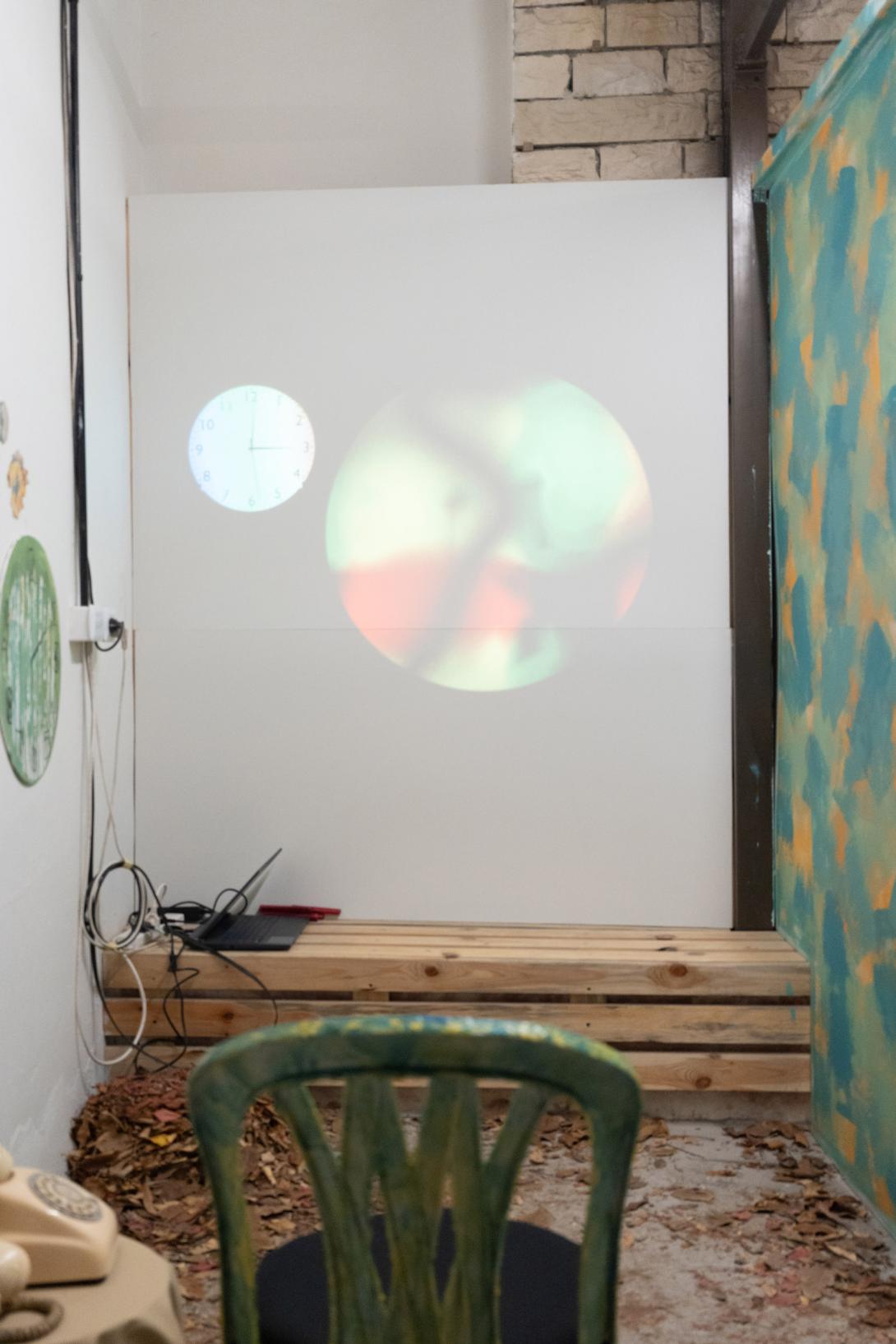 Low shot of the backside of a colorful plastic chair with hues of green,orange, and blue. Pointing at a wall with a clock projection and a circular projection with black, yellow, red, and green hues. On the ground is leafy foliage spread out.