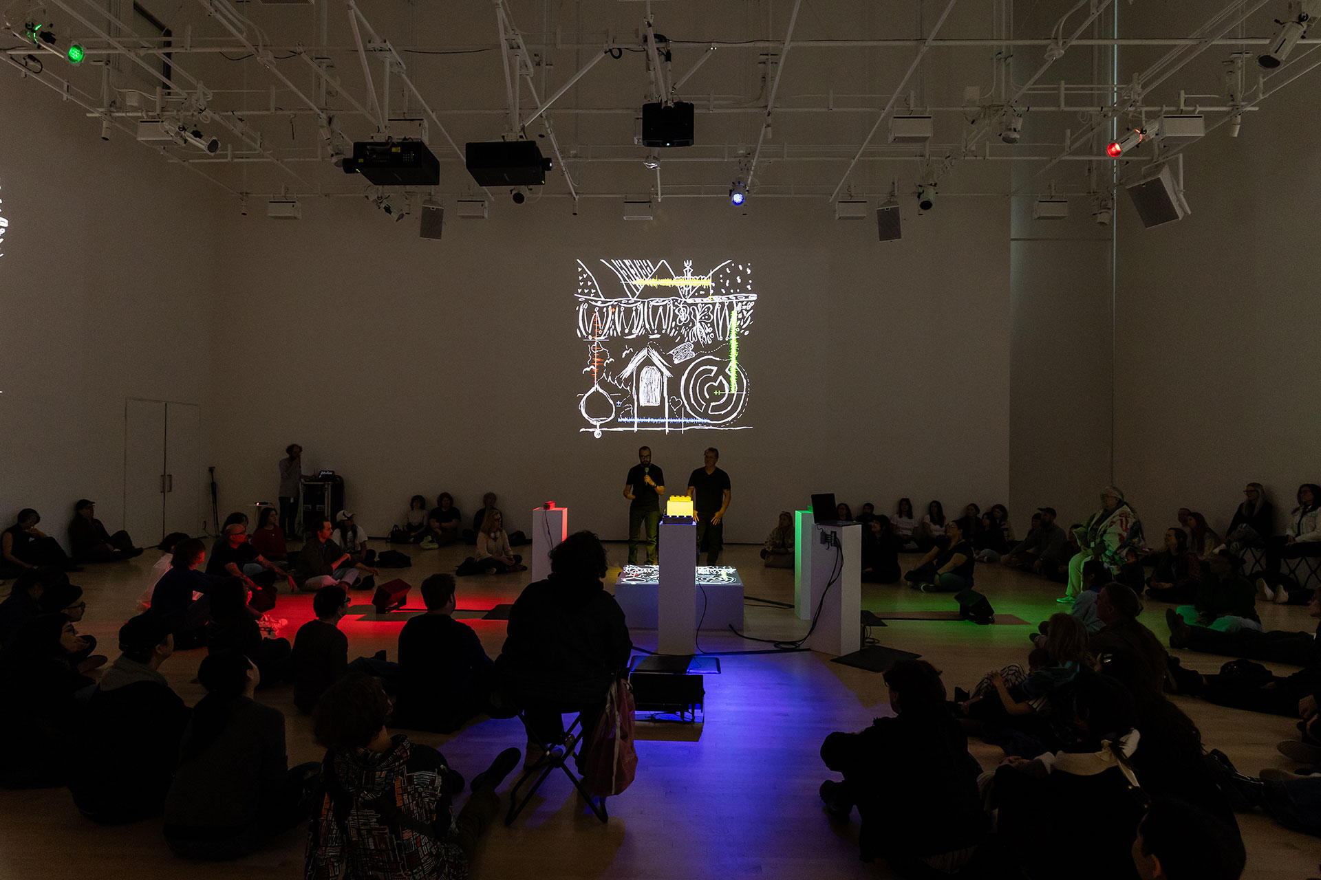 A dim, white walled room with an audience watching two people speak behind a circle of glowing podiums, with a white light projection of a sketchy illustration behind them.