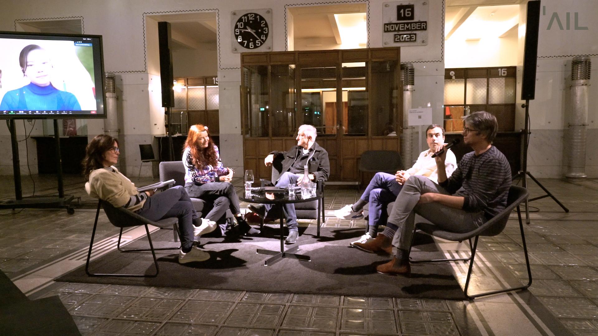 Five people sit on chairs in a half-circle. A screen behind them shows another person on videocall.