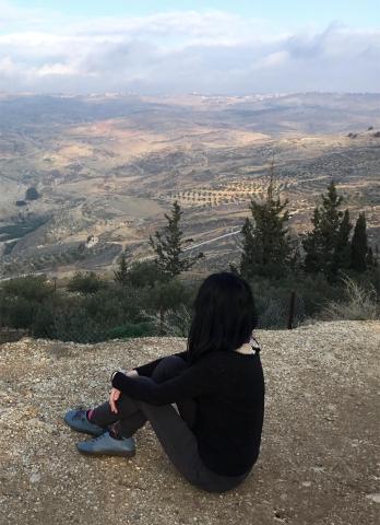 A woman with black hair seen from behind, wearing a black long-sleeved shirt, dark grey pants, and grey-blue shoes. She's sitting on the edge of a mountain overlooking a vast landscape of trees, paths, and hills. The sky beyond is blue with clouds on the horizon.