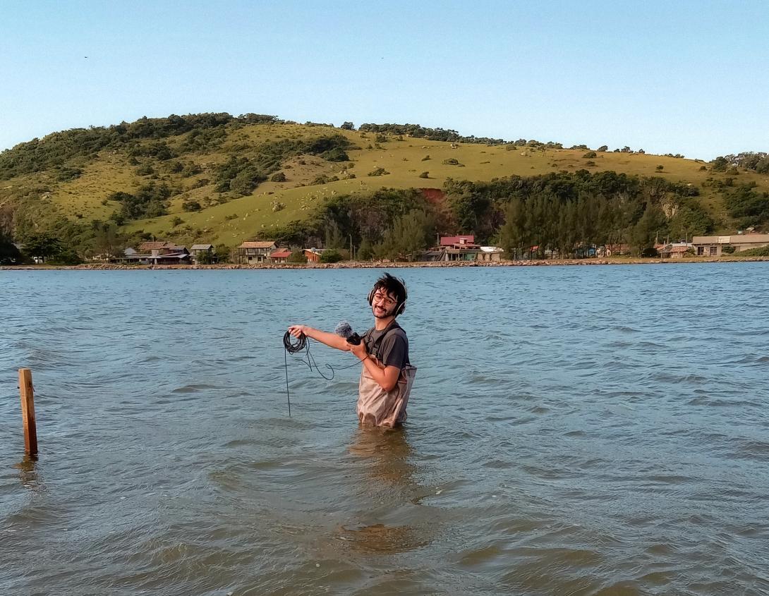 A smiling man with dark hair standing in a body of water, wearing headphones and holding up a wired microphone. In the background, houses line the edge of the water, and beyond are rolling green hills dotted with trees and a blue, cloudless sky.