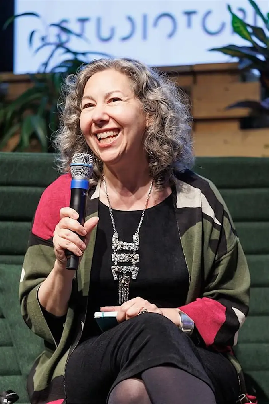Woman with grey, wavy hair smiling while holding up a microphone. She's sitting down on a green fabric chair, wearing a black dress and a green cardigan with a red, black, and white geometric design. An out-of-focus screen and plants are visible in the background.